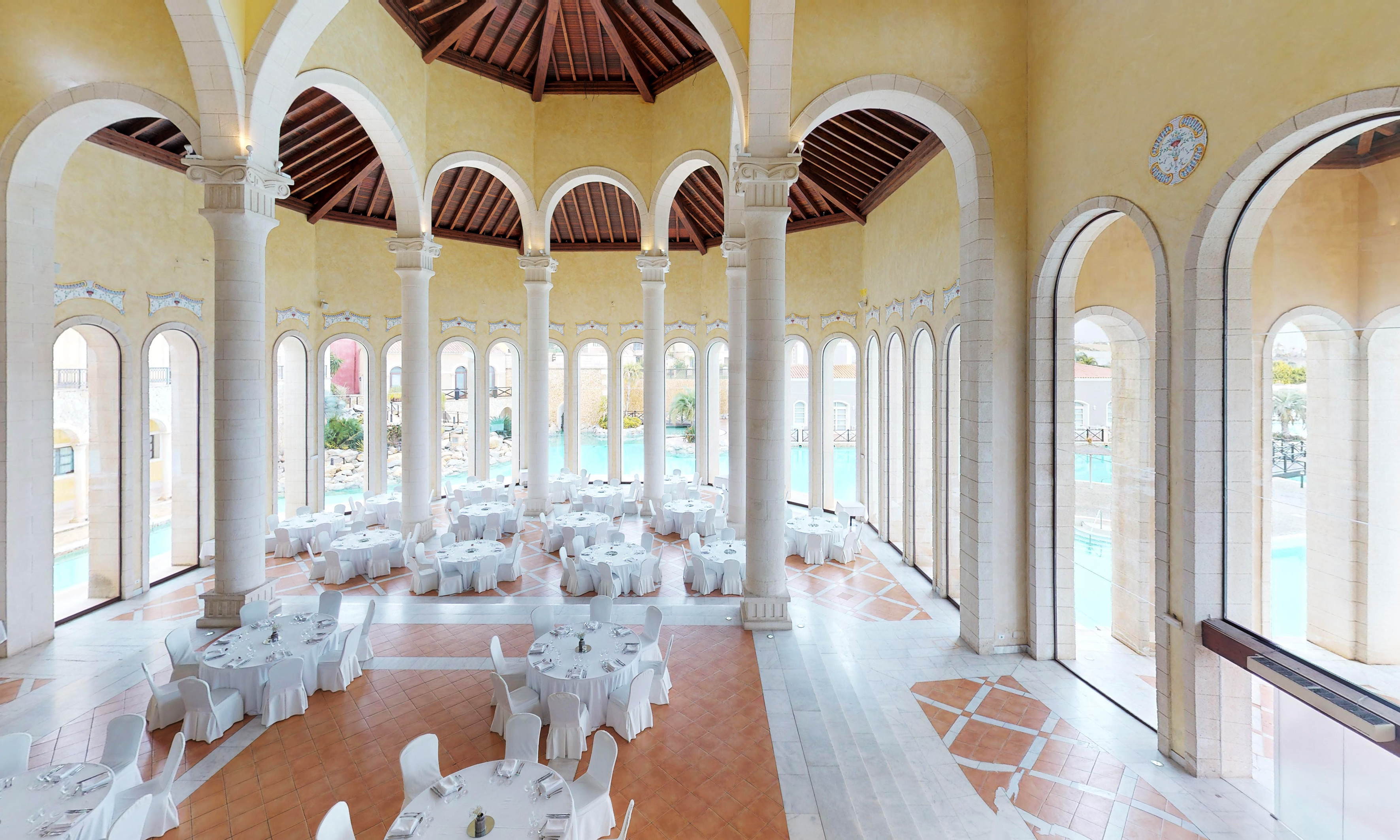 Elegant Mediterranean hall with white tables, columns, overlooking resort pool.