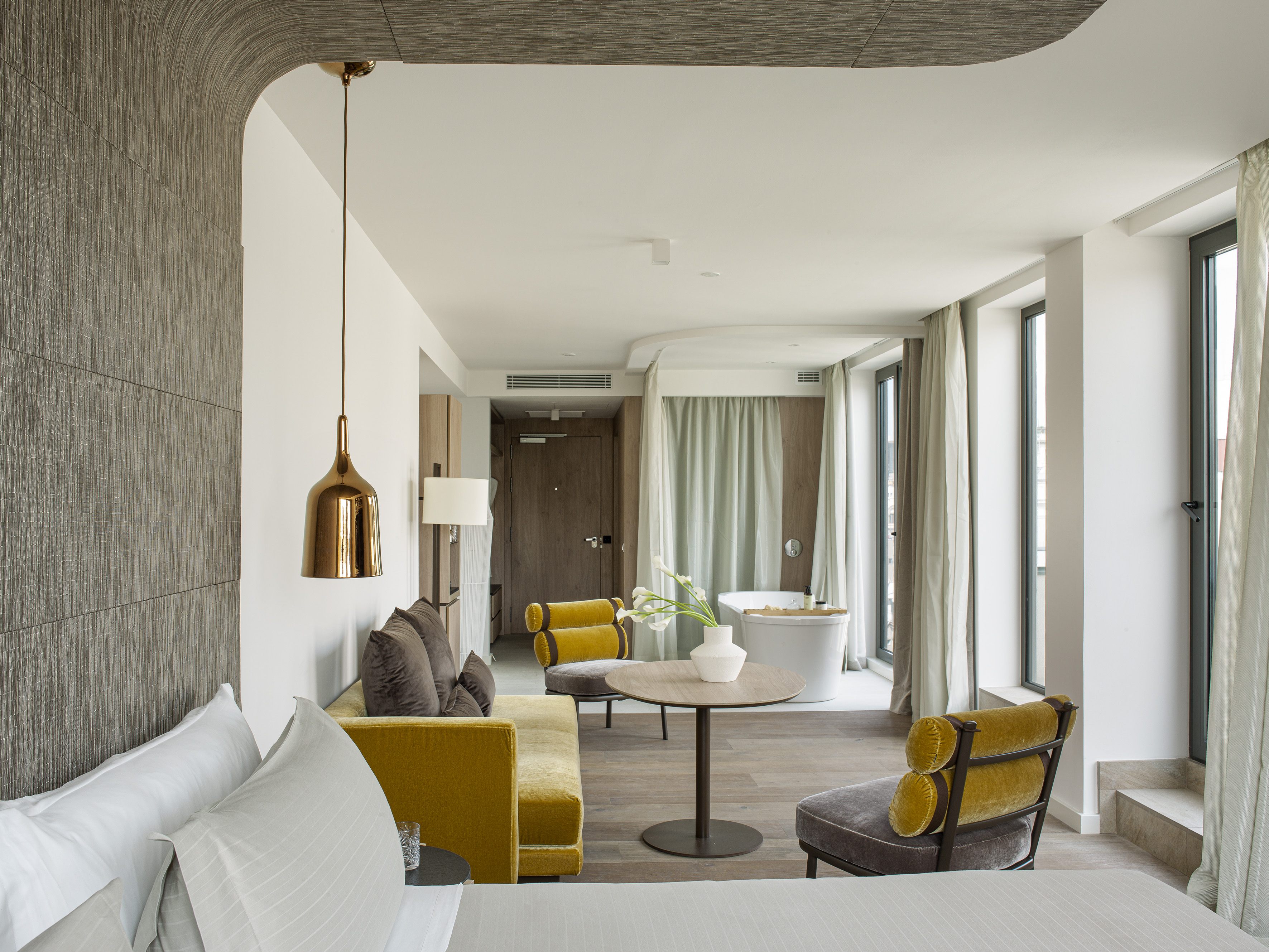 Modern hotel suite with yellow sofa, freestanding bathtub, and large windows.