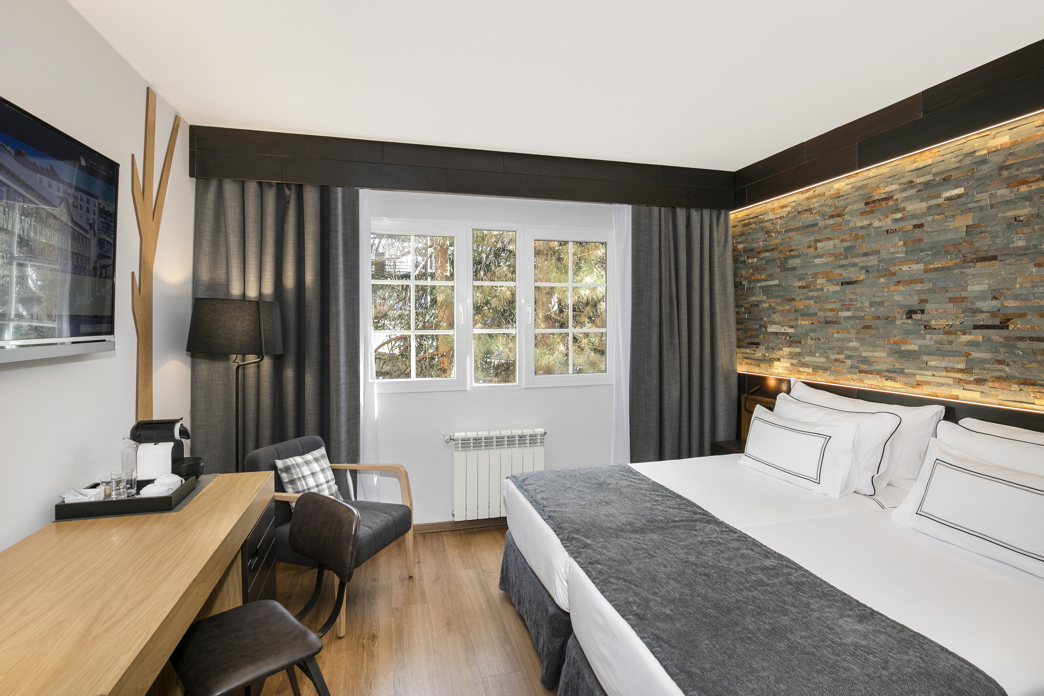 Modern hotel bedroom: large bed, stone accent wall, window, desk, coffee machine.