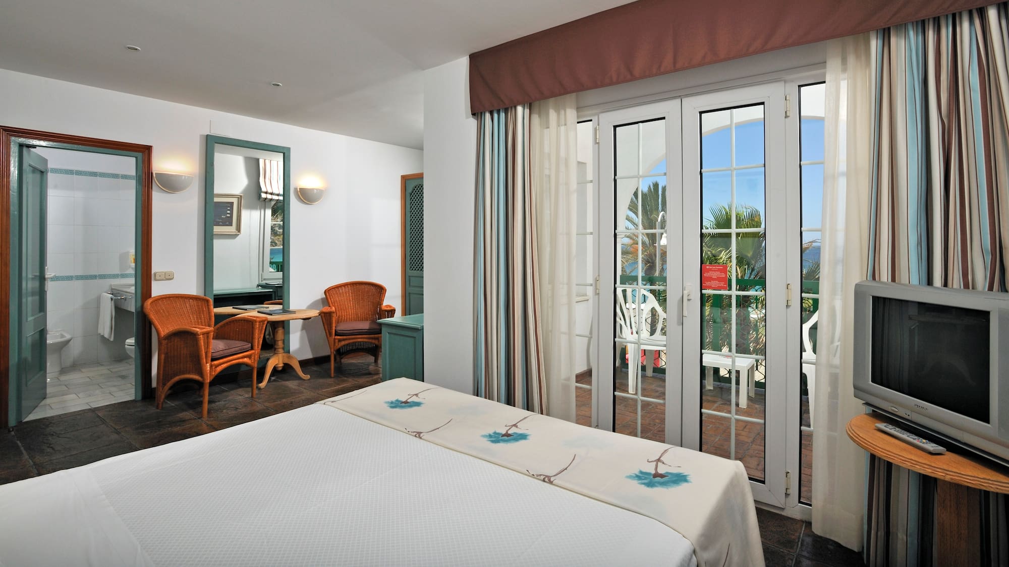 Hotel bedroom with ocean-view balcony, white bed, and rattan chairs.