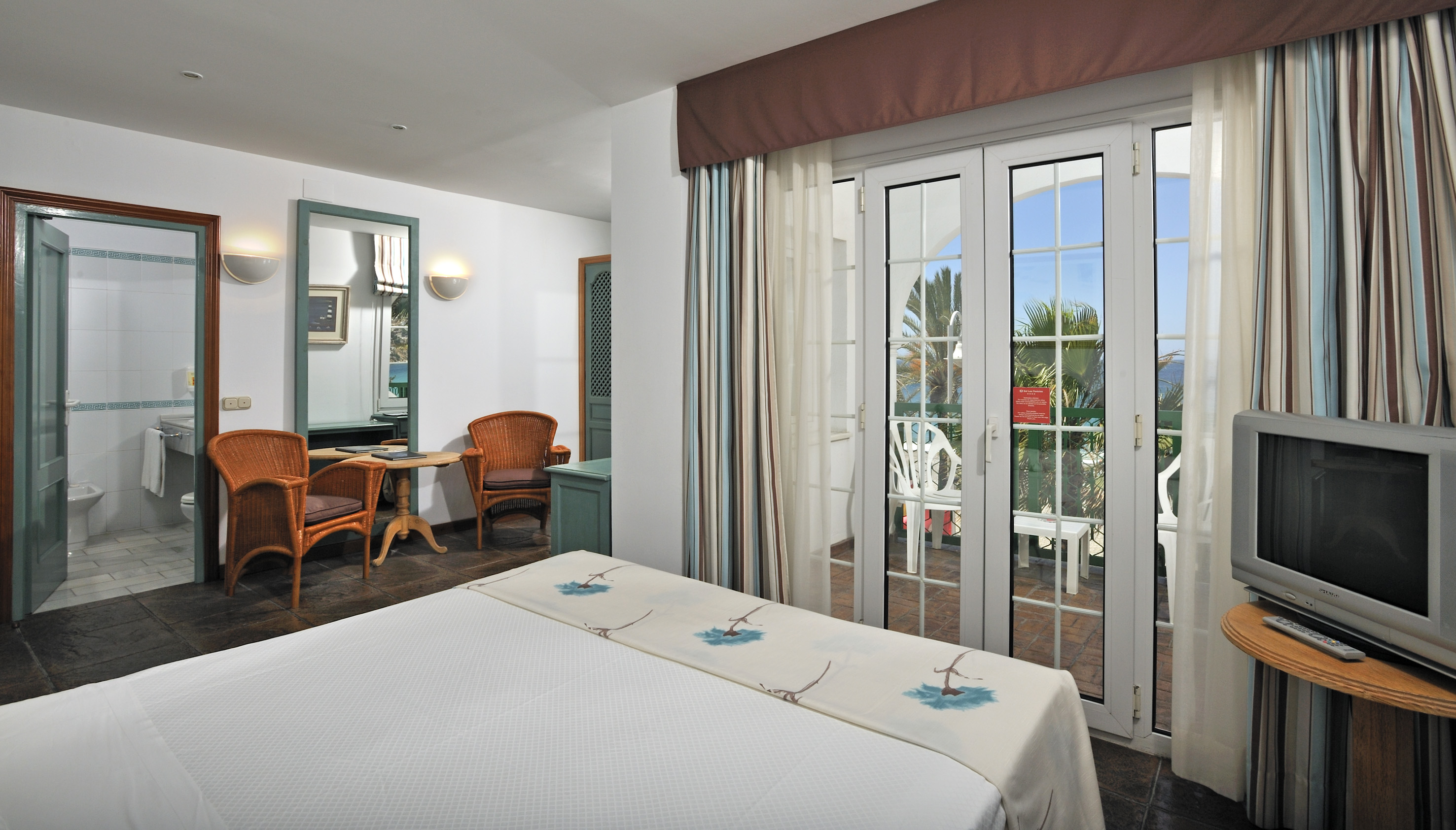 Hotel bedroom with ocean-view balcony, white bed, and rattan chairs.