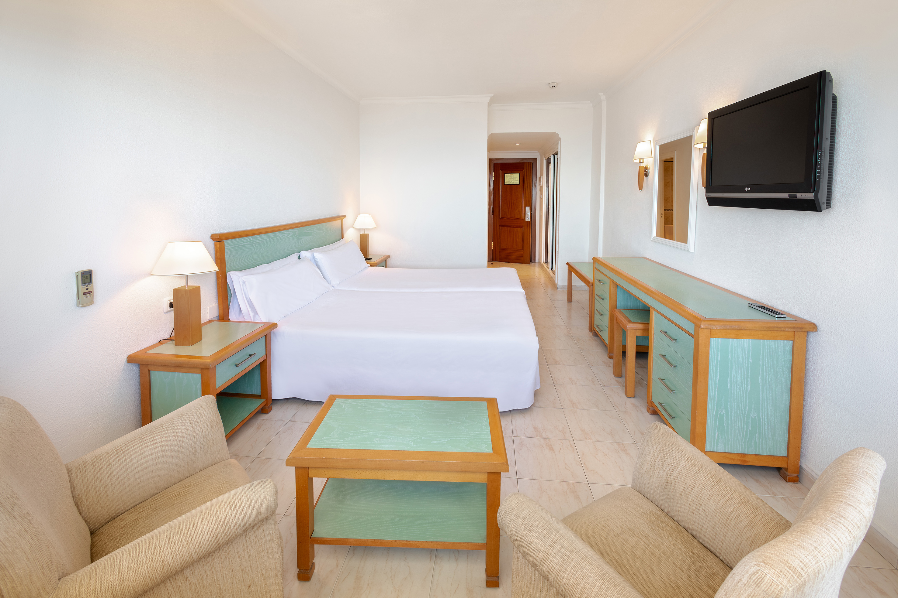 Hotel room with two beds, wood & teal furniture, wall-mounted TV, armchairs.
