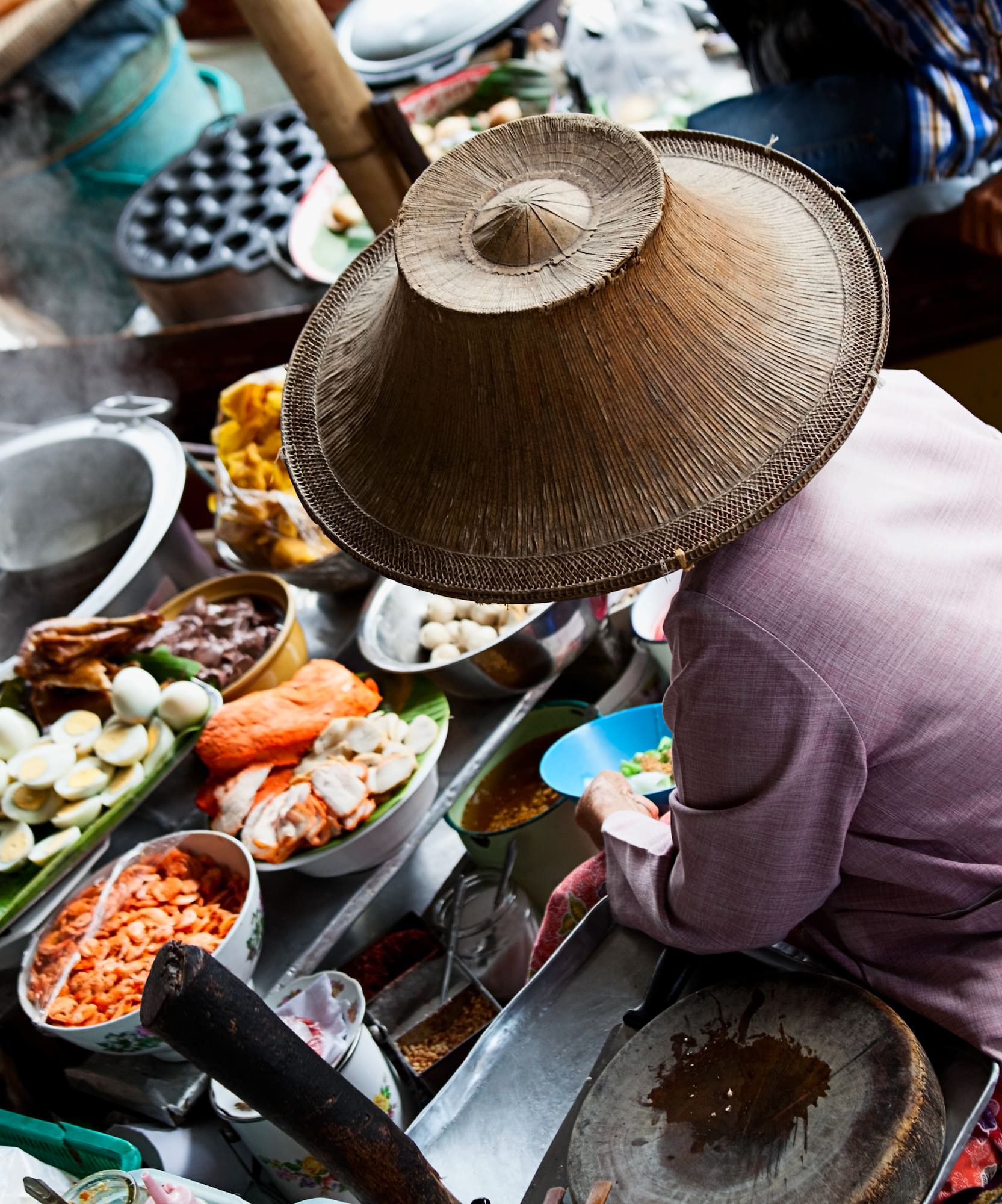 Thai floating market vendor in conical hat prepares food on boat.