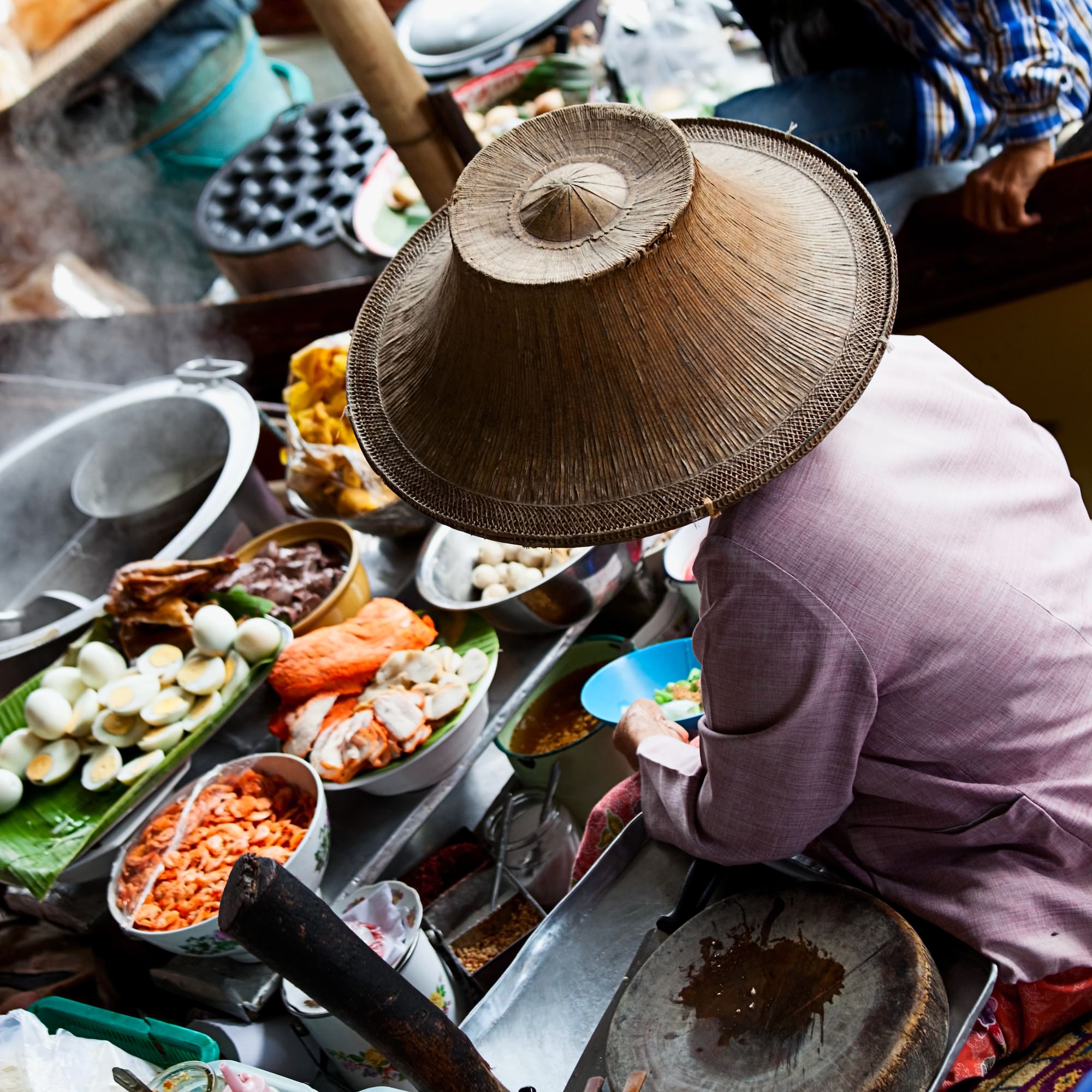 Thai floating market vendor in conical hat prepares food on boat.