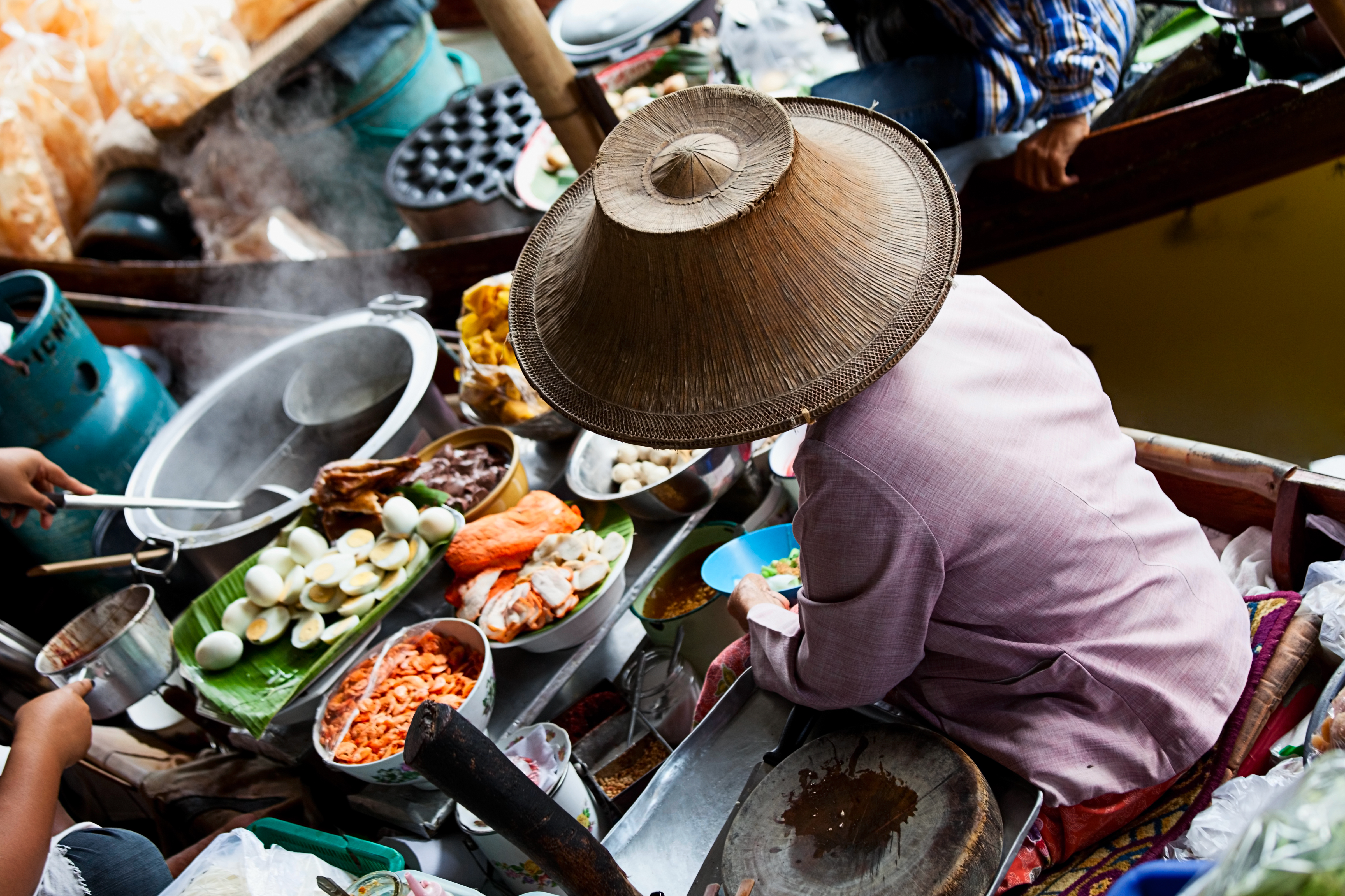 Thai floating market vendor in conical hat prepares food on boat.