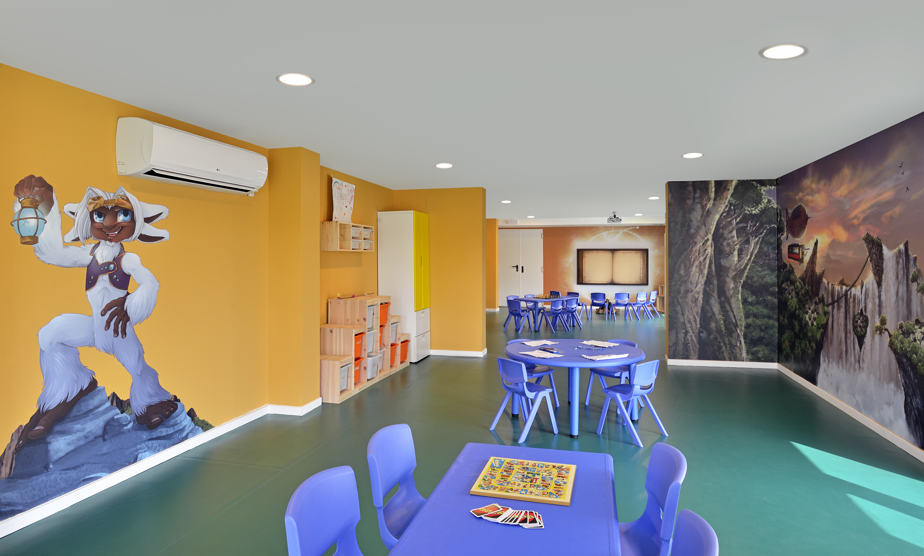 Bright kids' club room featuring cartoon murals, purple tables, and games.