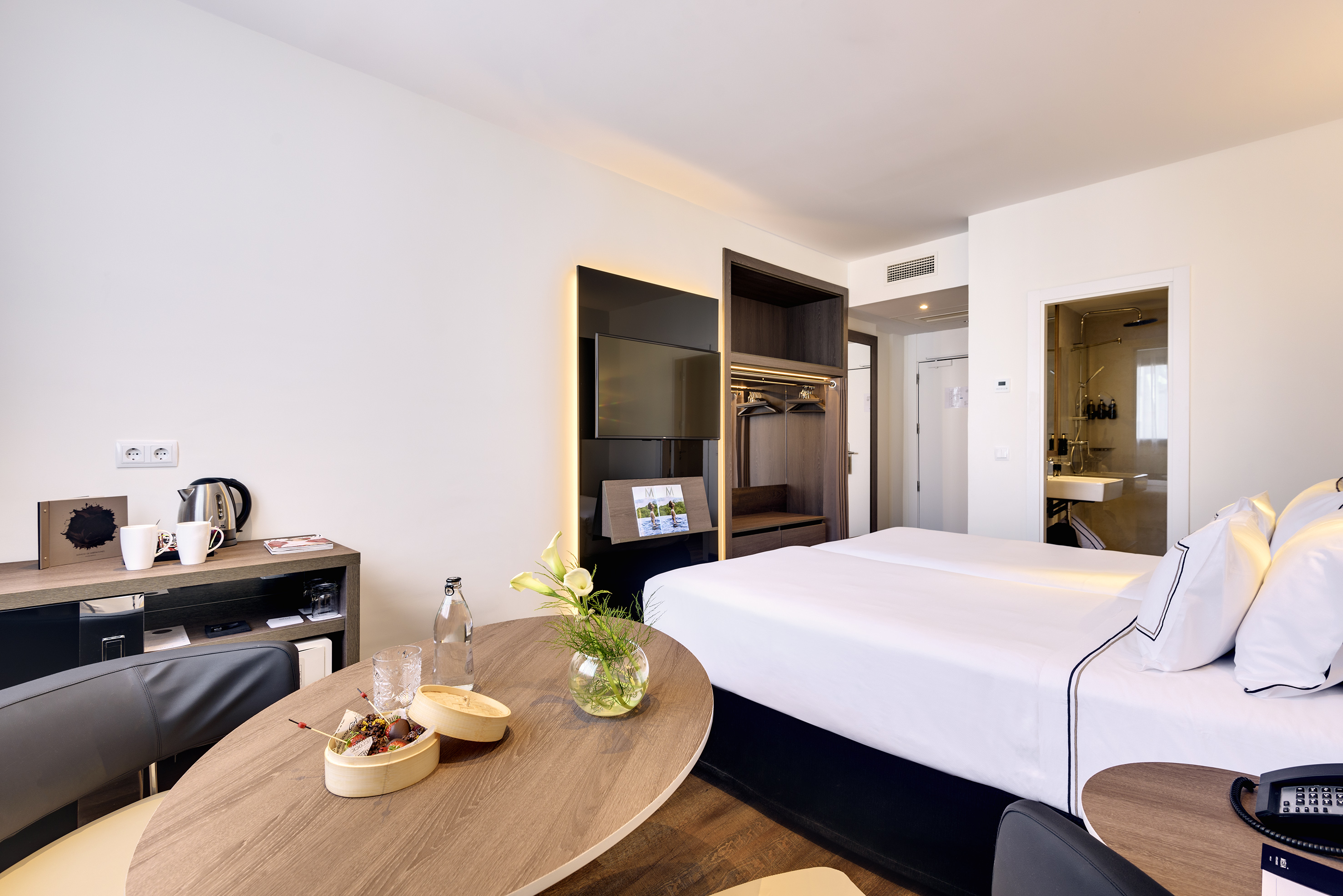 Modern hotel room with two beds, snack table, TV, and open wardrobe.