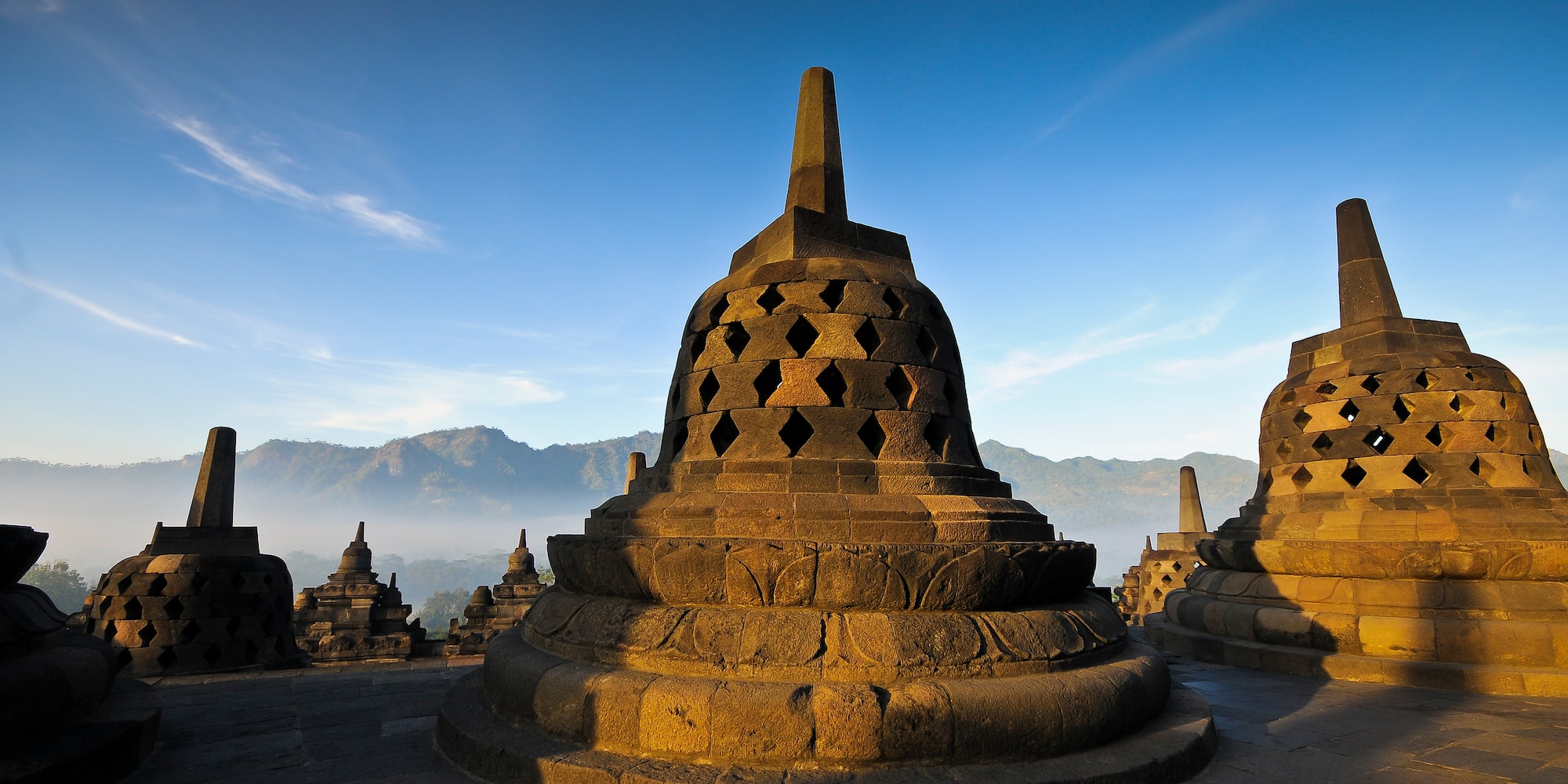 Sunlit Borobudur temple stupas with misty mountains at sunrise.