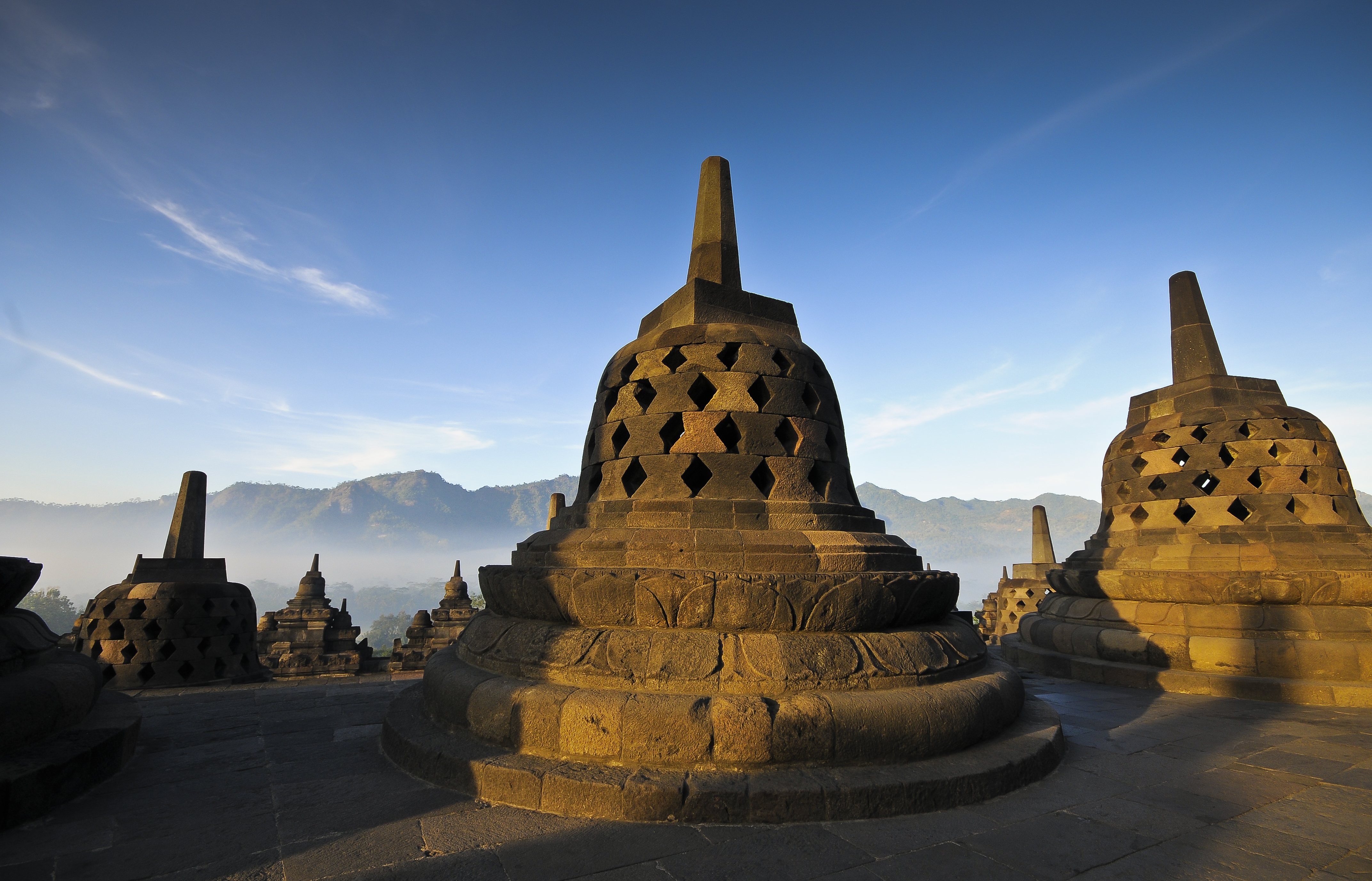 Sunlit Borobudur temple stupas with misty mountains at sunrise.