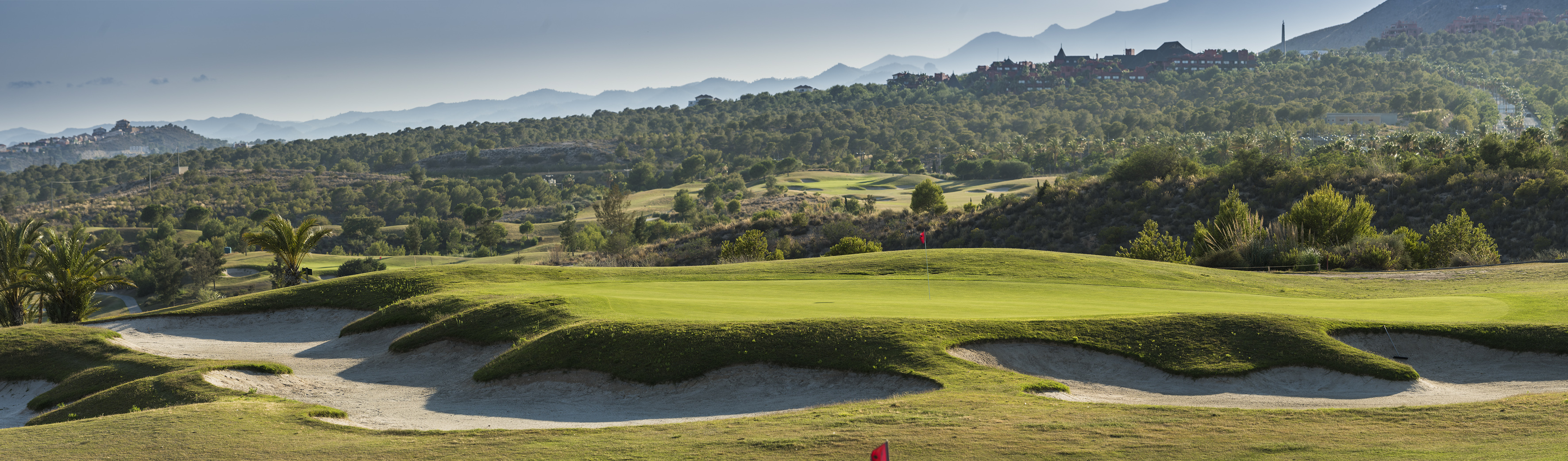 Lush green golf course with sand bunkers, mountains, and a distant resort building.