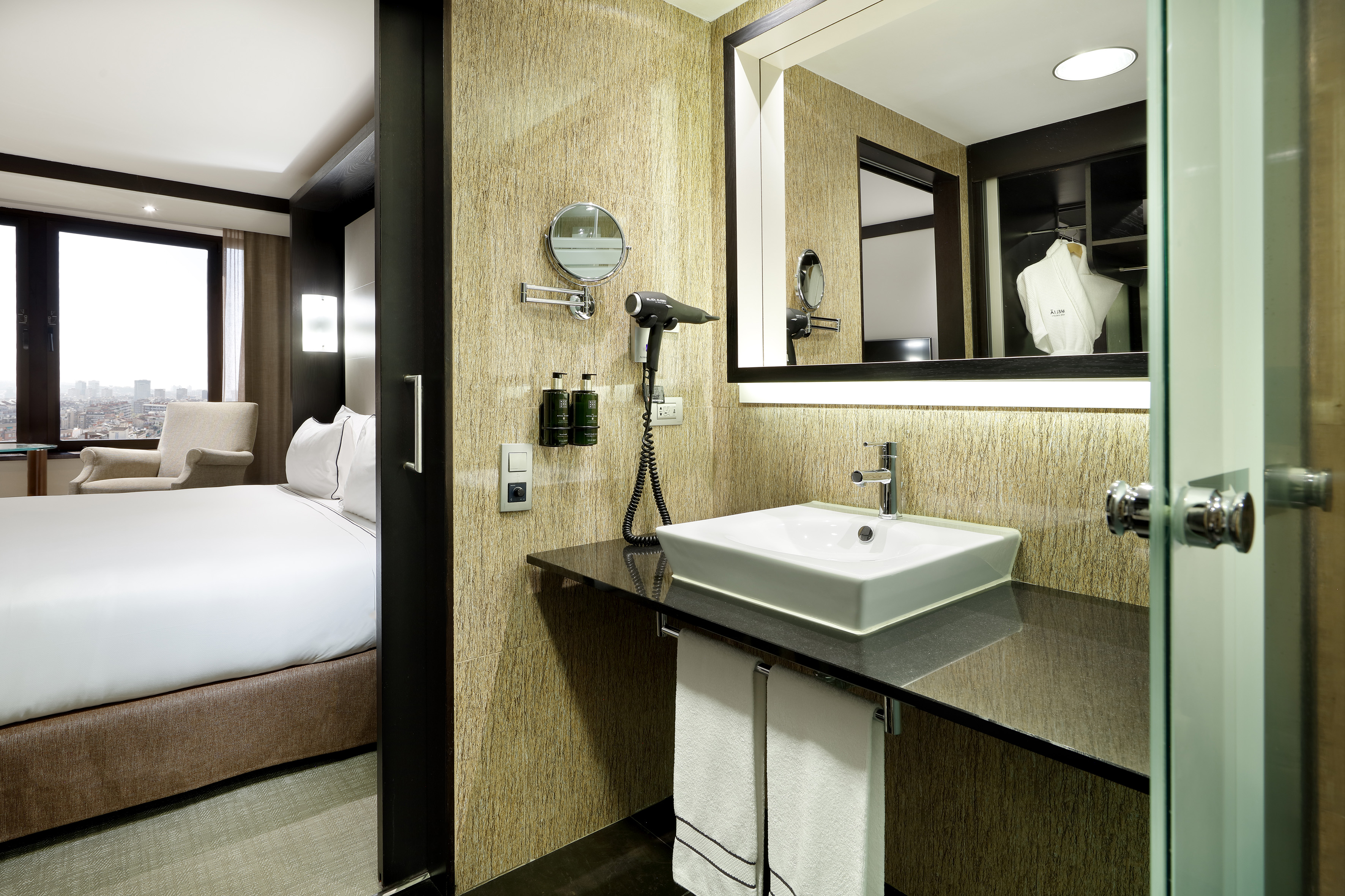 Hotel room: modern bathroom vanity, sink, mirror, bed, and city view.
