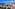 Coastal resort: large blue pool, hotels, palm trees, and ocean view.