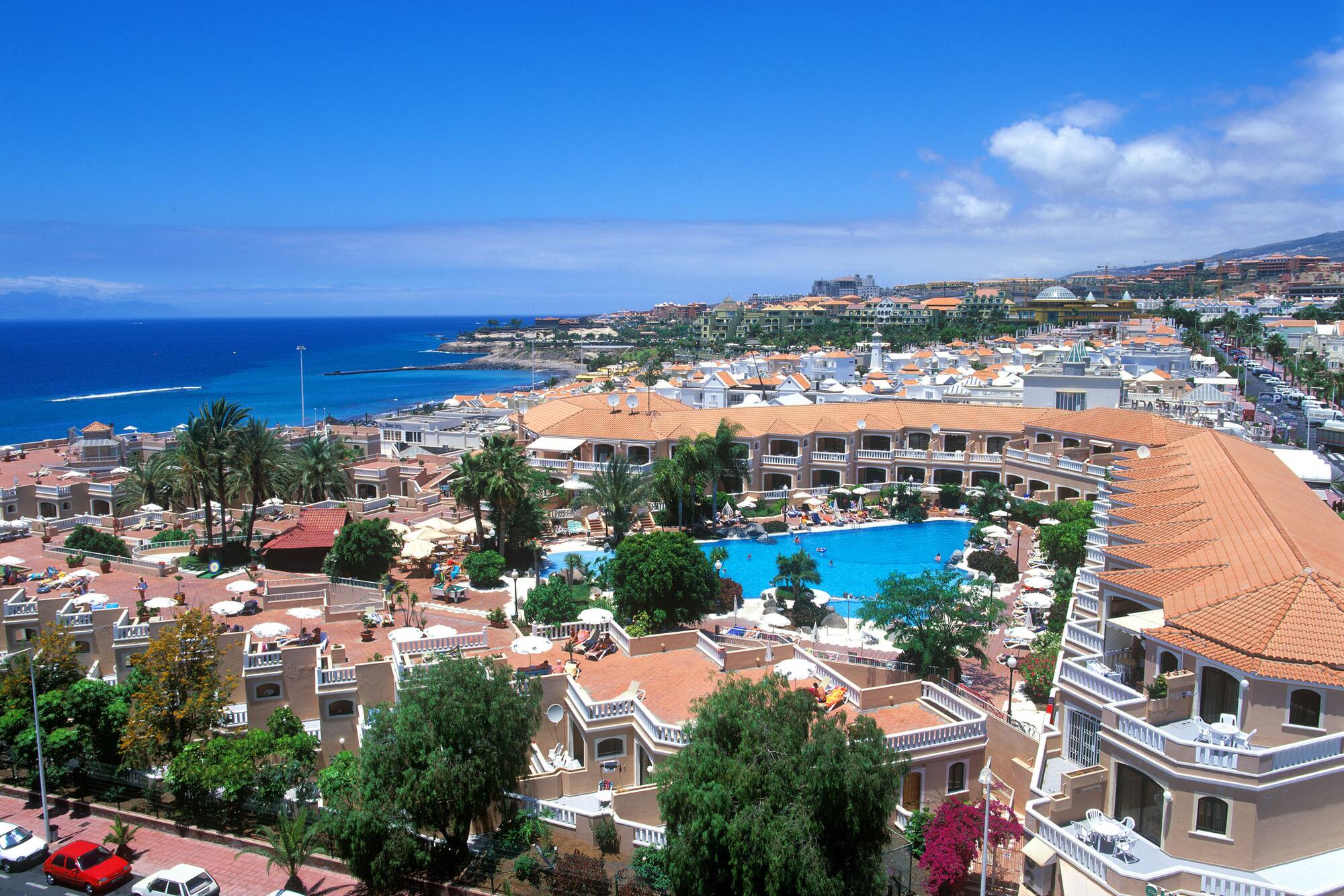 Coastal resort: large blue pool, hotels, palm trees, and ocean view.