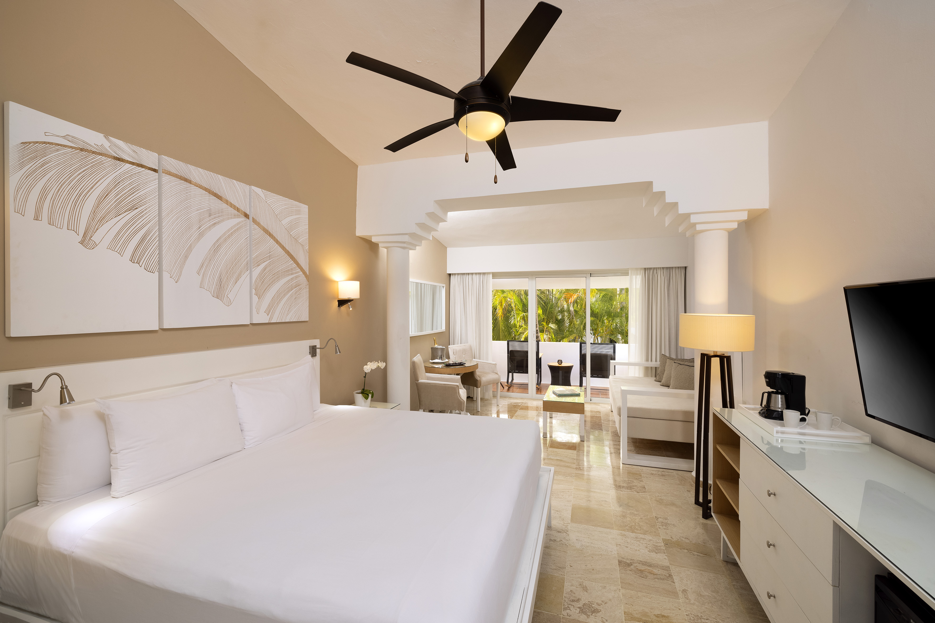 Modern hotel room with king bed, living area, and tropical balcony view.
