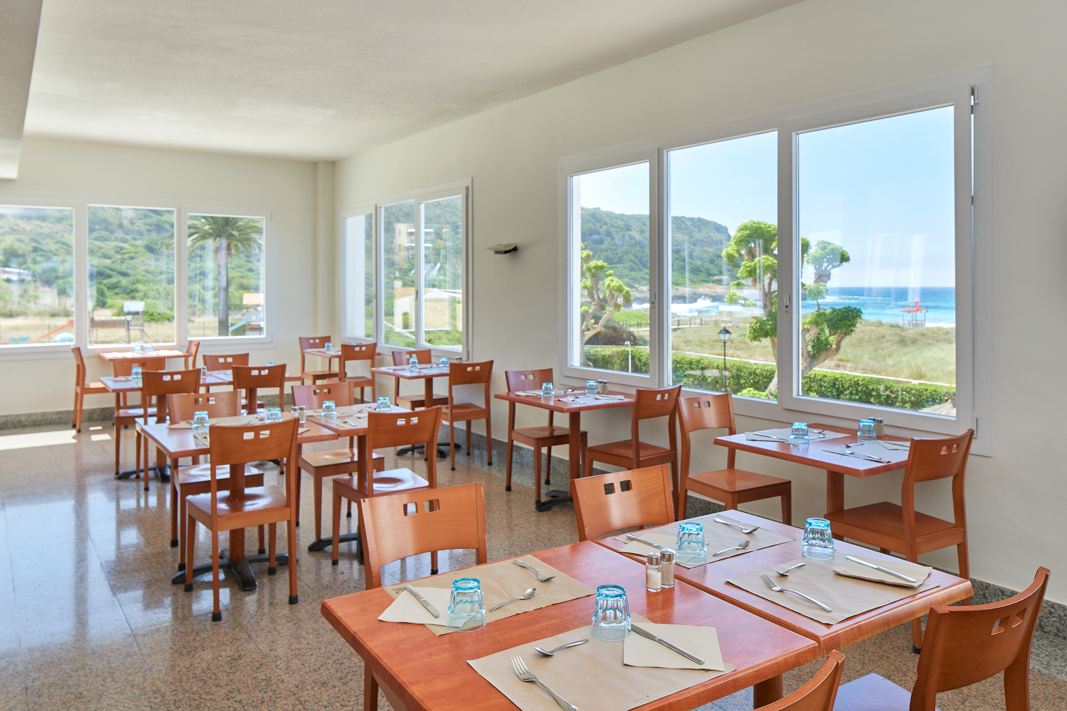Resort dining room with set tables and ocean beach view through large windows.