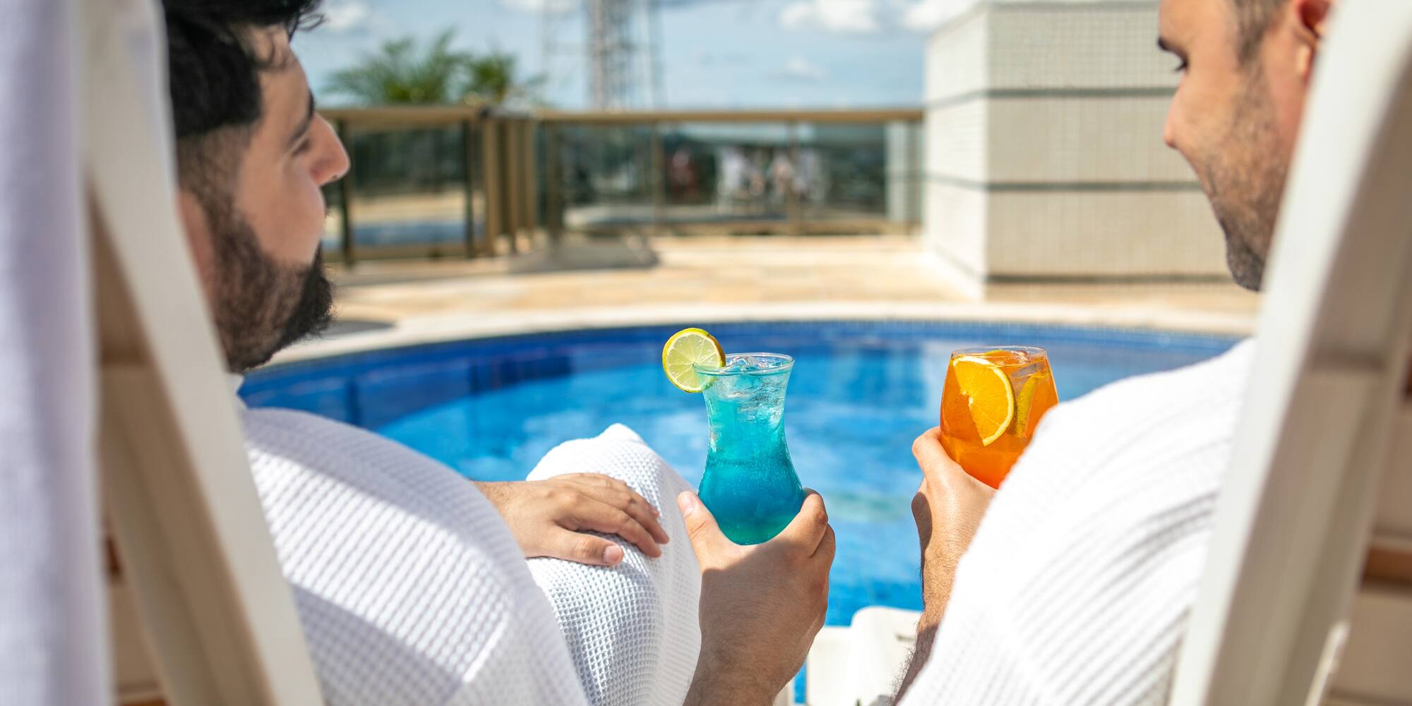 two men sitting in chairs holding drinks by a pool
