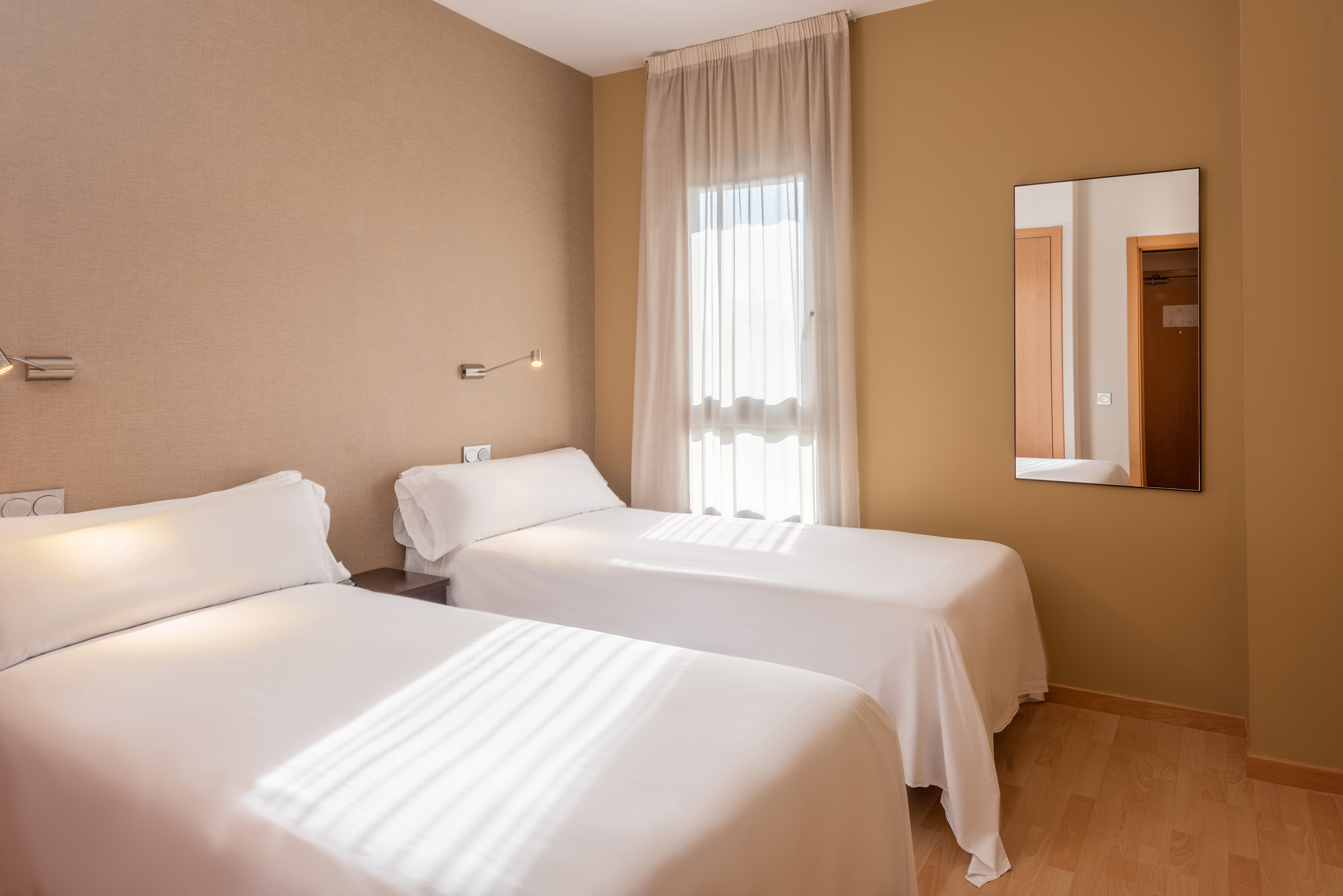 Hotel room with two single beds, beige walls, and sheer window curtains.