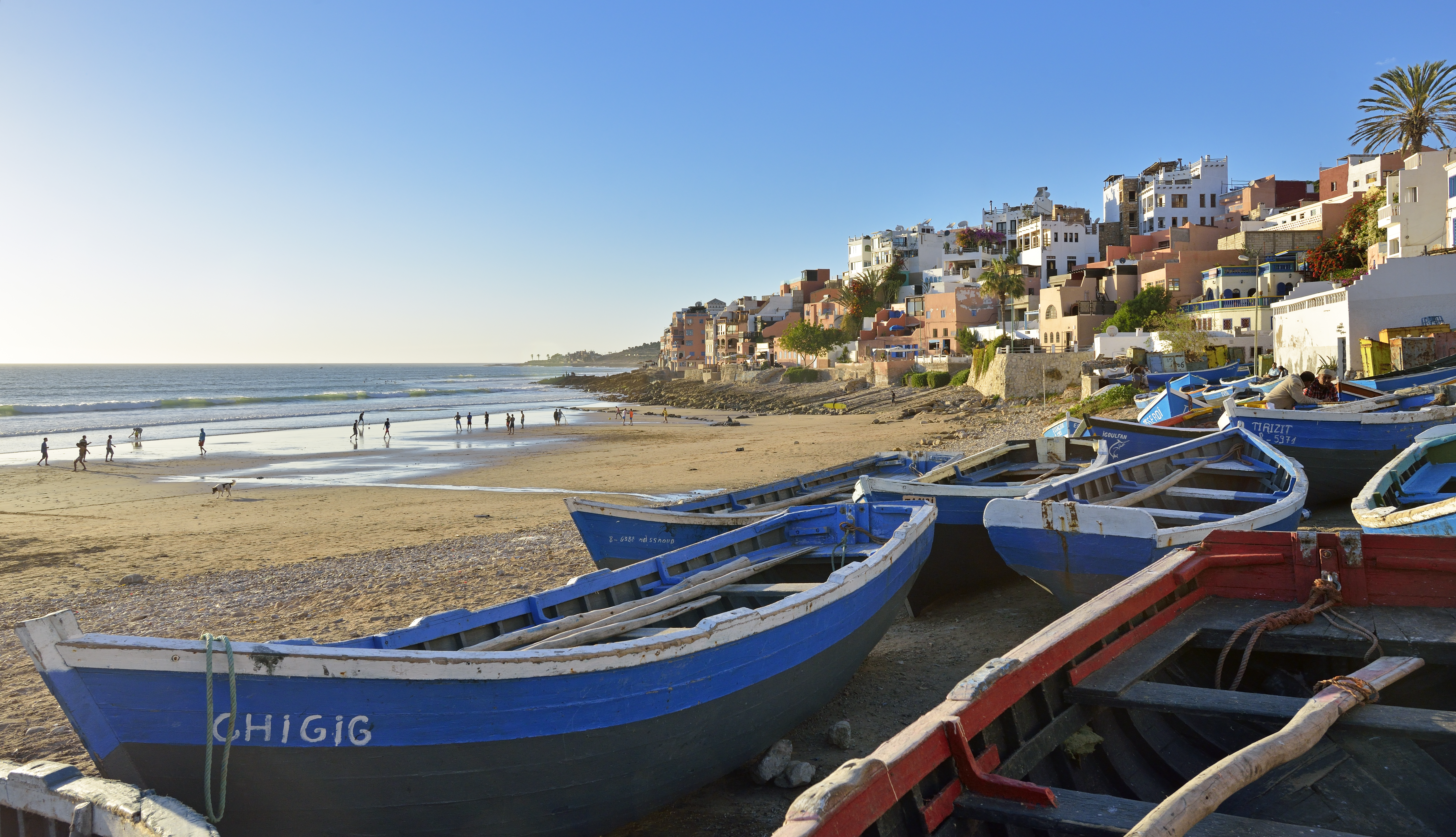 Blue fishing boats on a sandy beach fronting a colorful cliffside town and ocean.