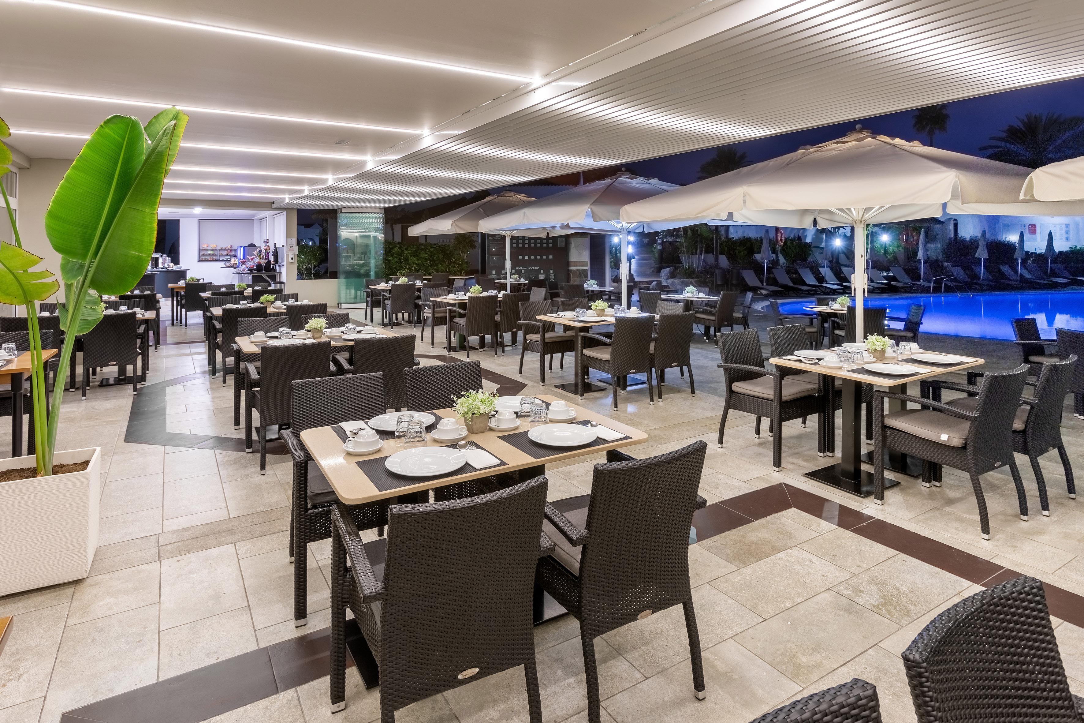Resort restaurant patio at night with set tables, pool, and umbrellas.
