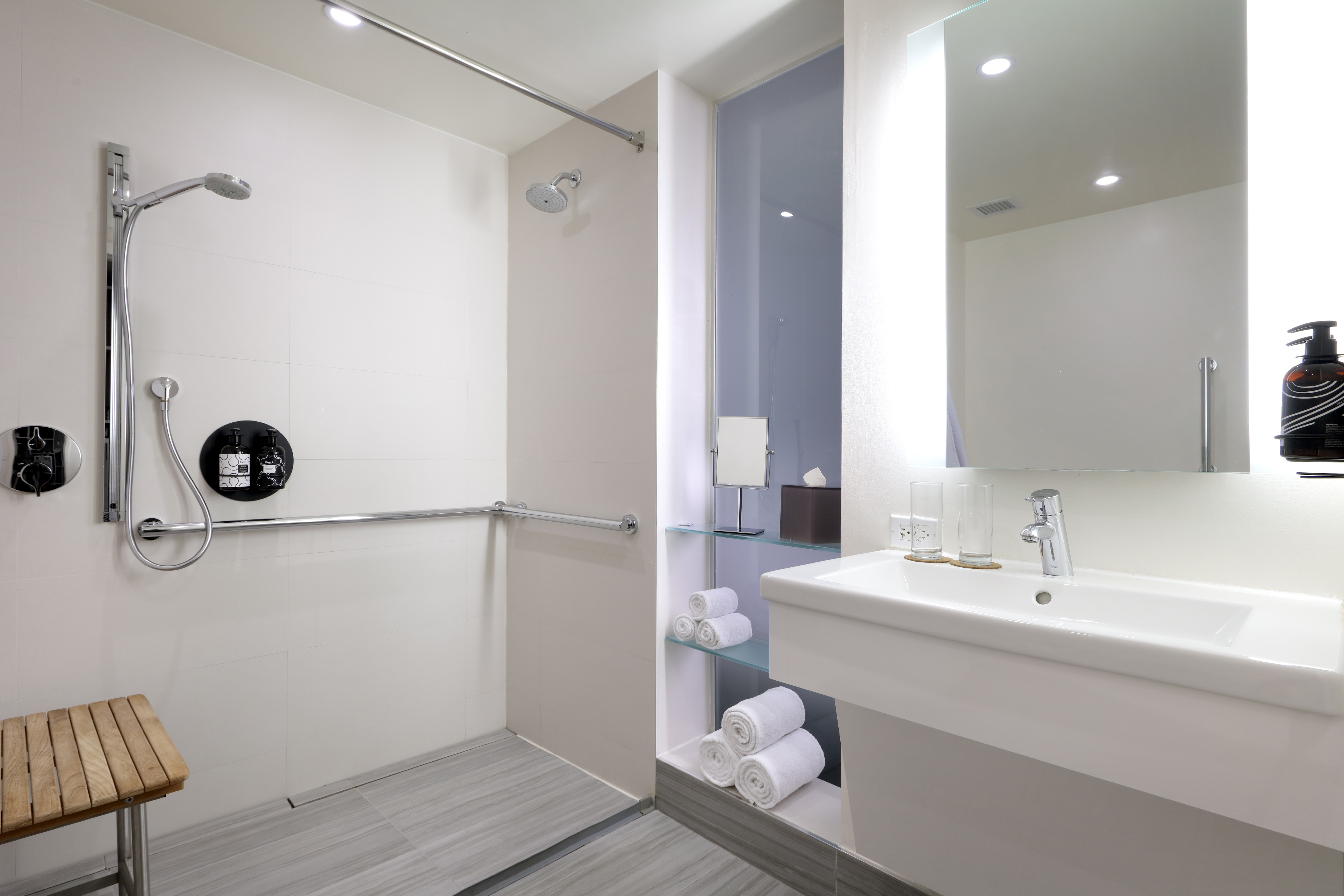 Modern accessible bathroom with grab bar, shower, wooden bench, and vanity.