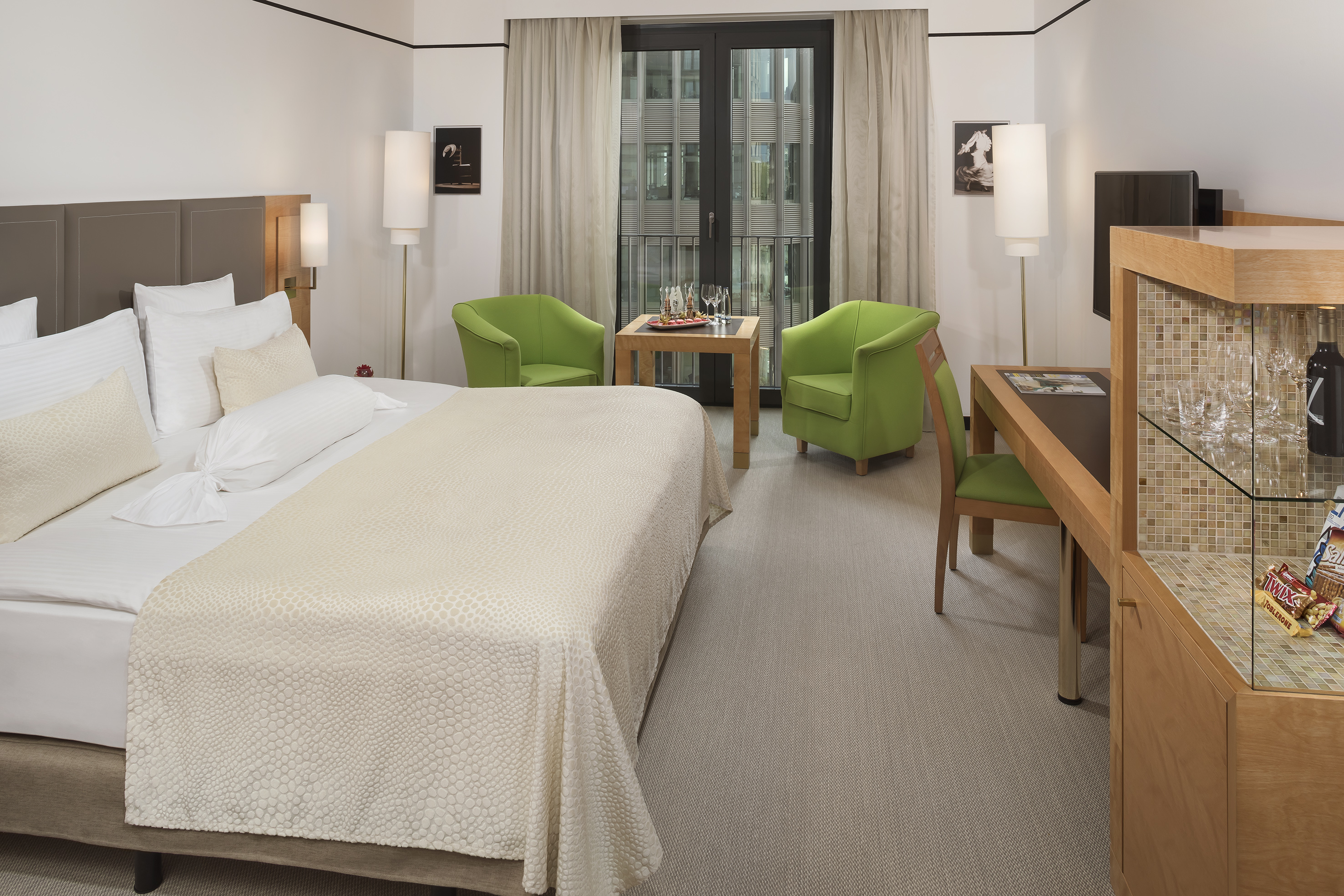 Modern hotel room with king bed, green armchairs, desk, and city view from balcony.