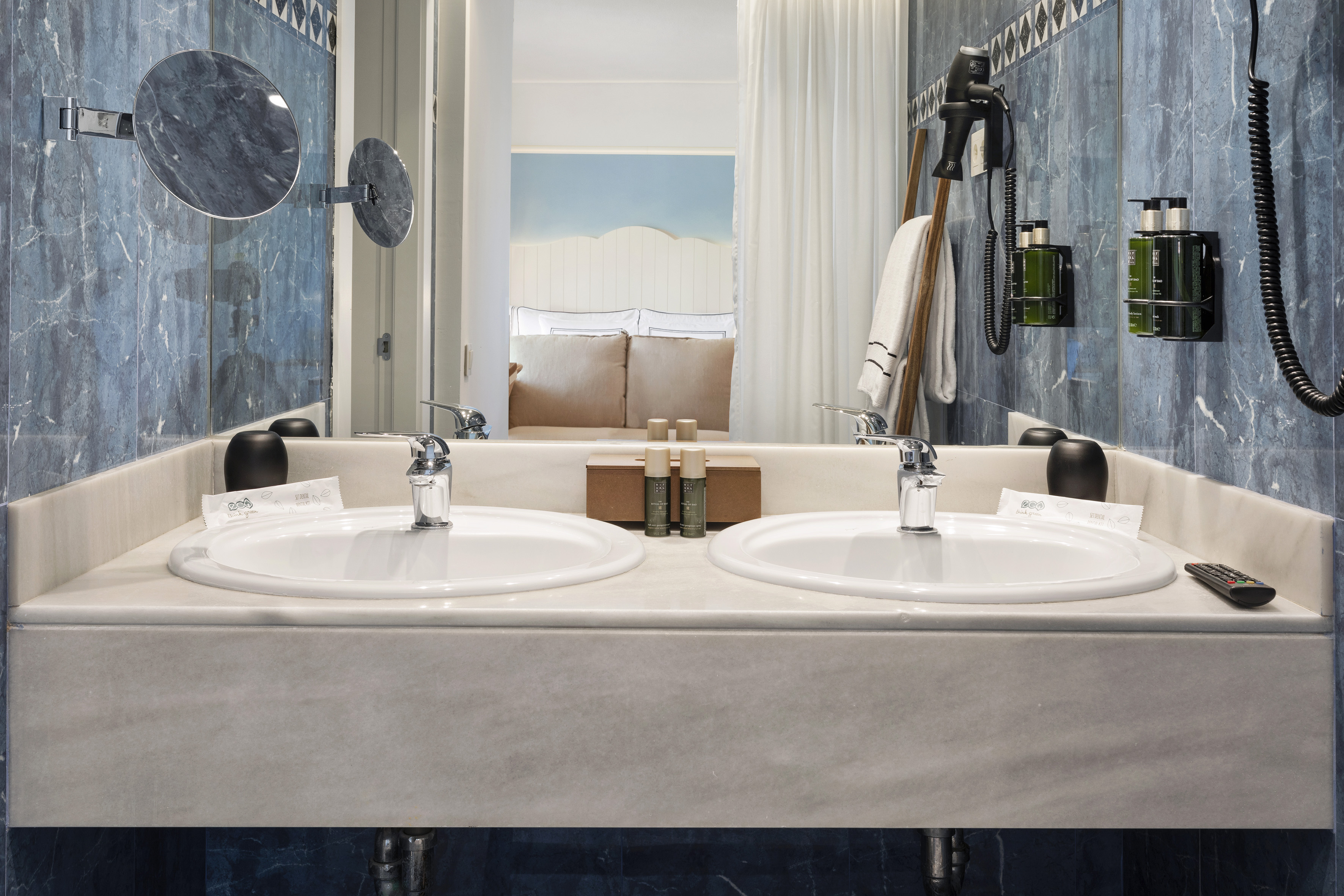 Hotel bathroom with double marble vanity, sinks, and mirror reflecting a bedroom.