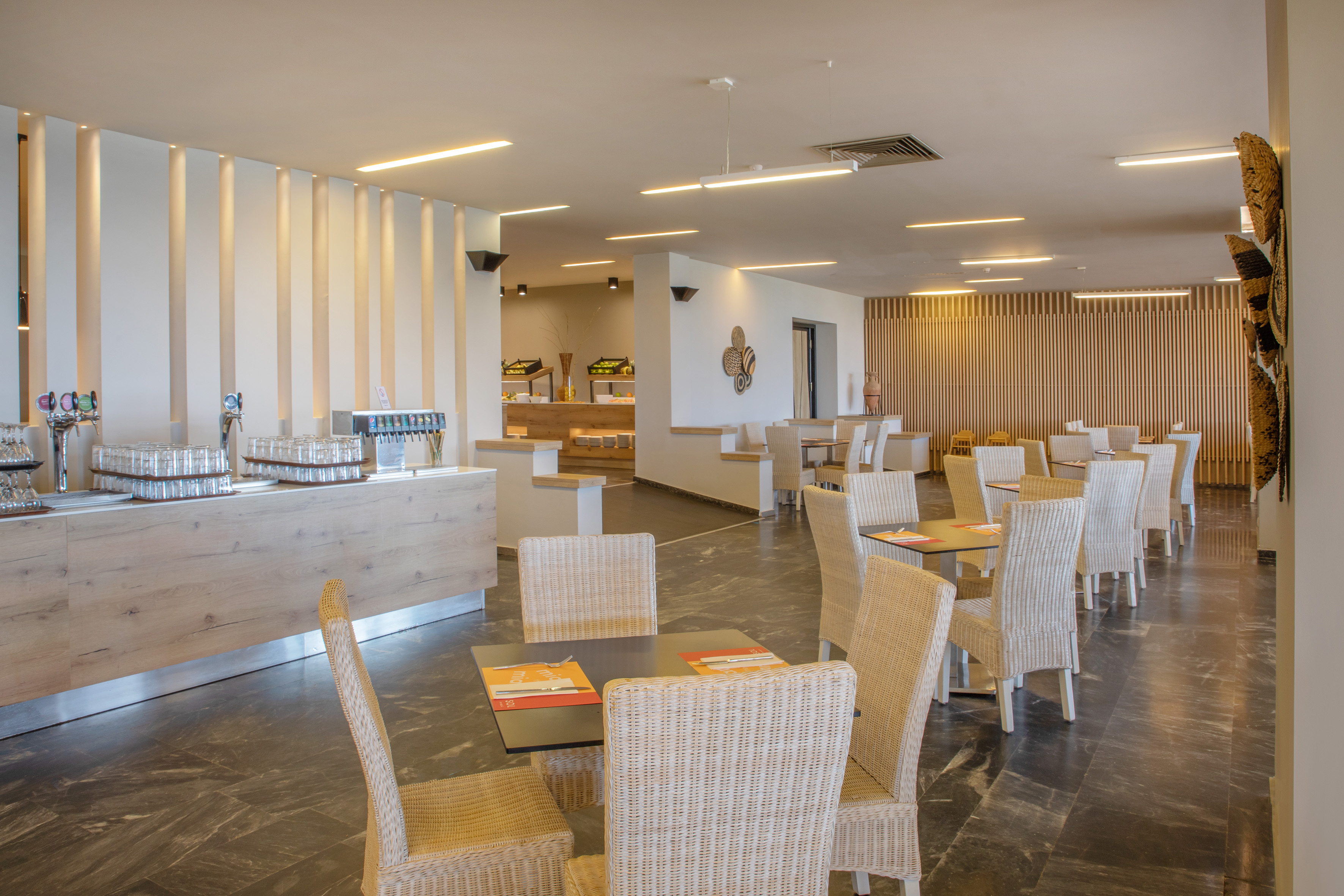 Modern hotel restaurant with buffet, wicker chairs, and dark tables.