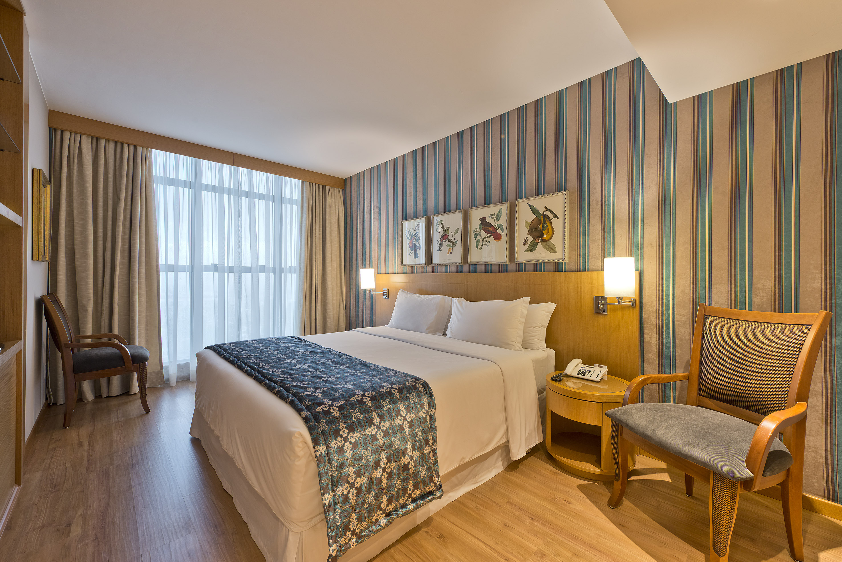 Stylish hotel room with king bed, striped wallpaper, wooden floor, and bright window.