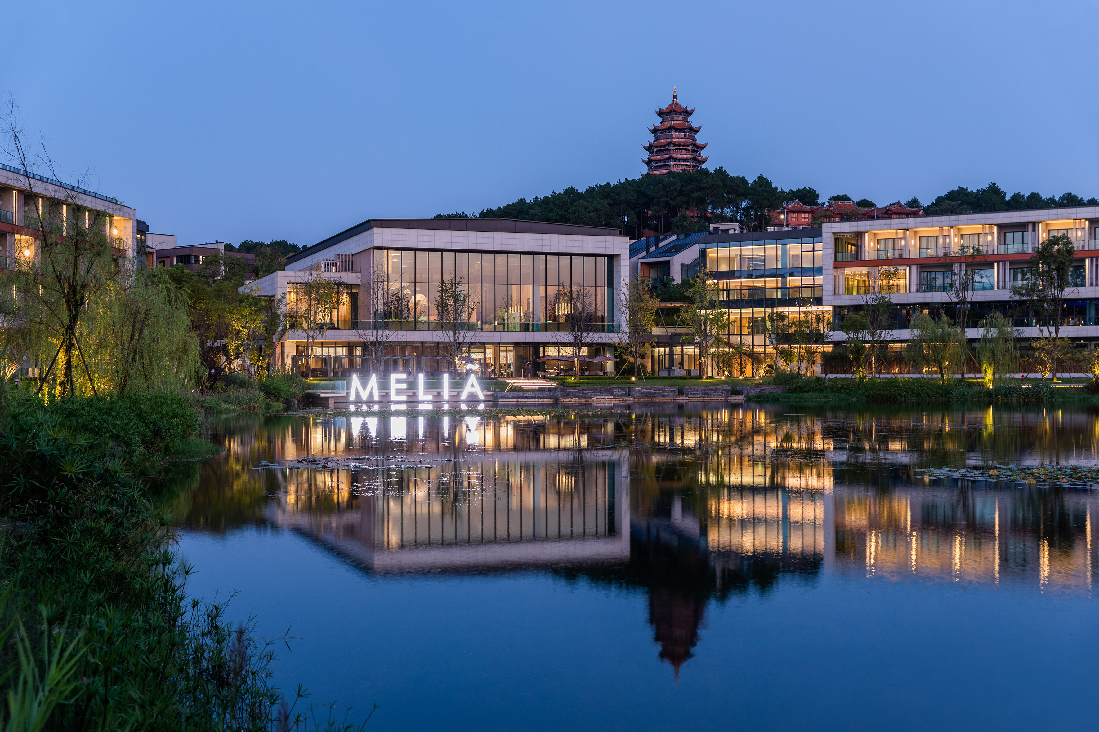 Illuminated Melia resort reflected in lake, with pagoda on hillside at dusk.
