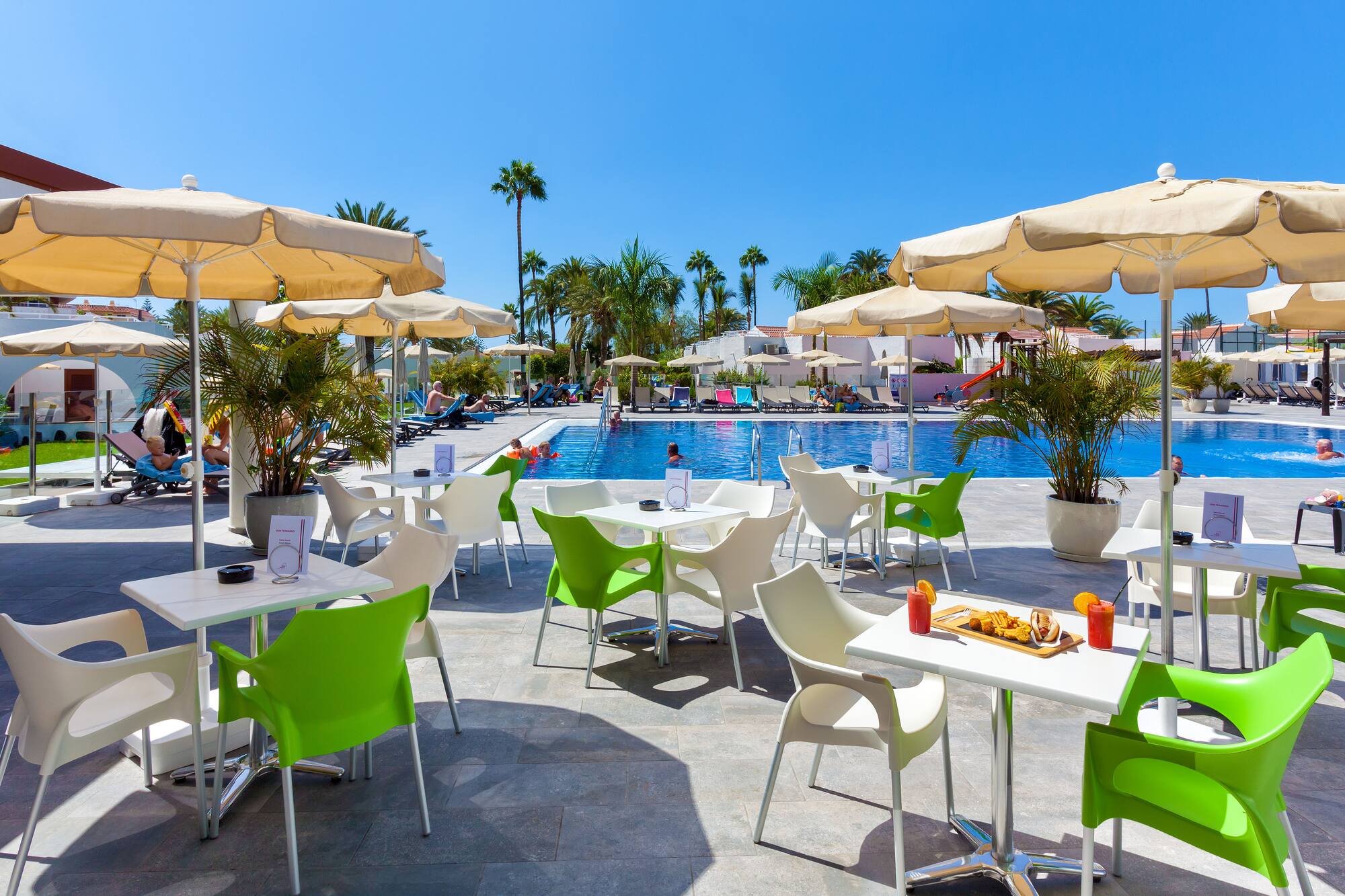 Sunny resort pool cafe patio with green chairs, tables, and lush palm trees.