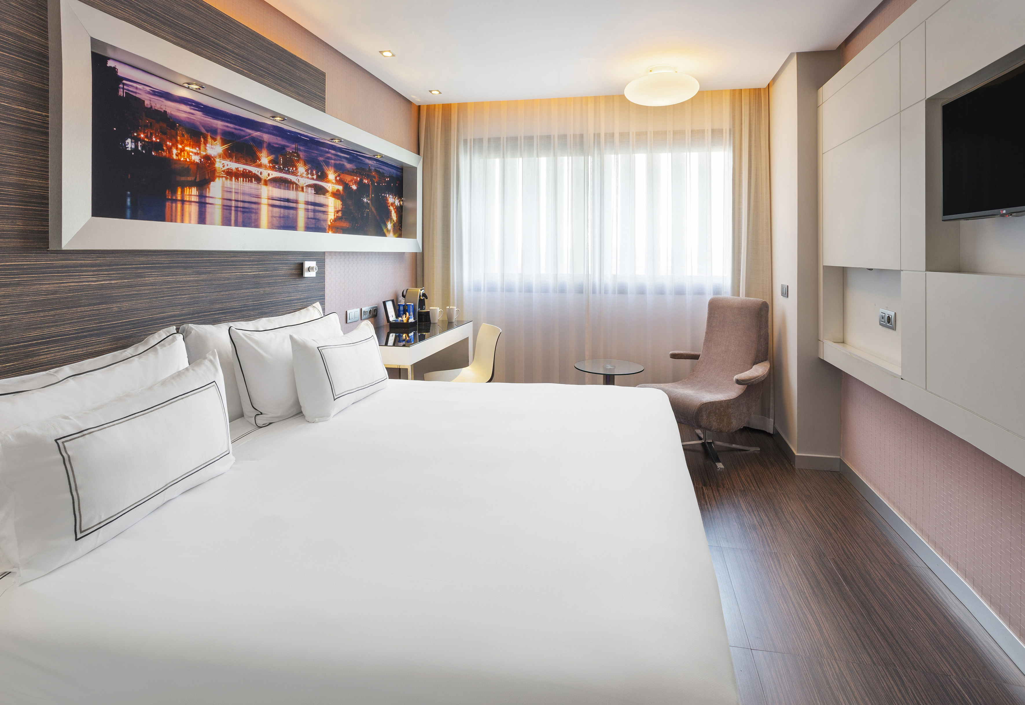 Spacious modern hotel room with white bed, city view art, desk, and chair.