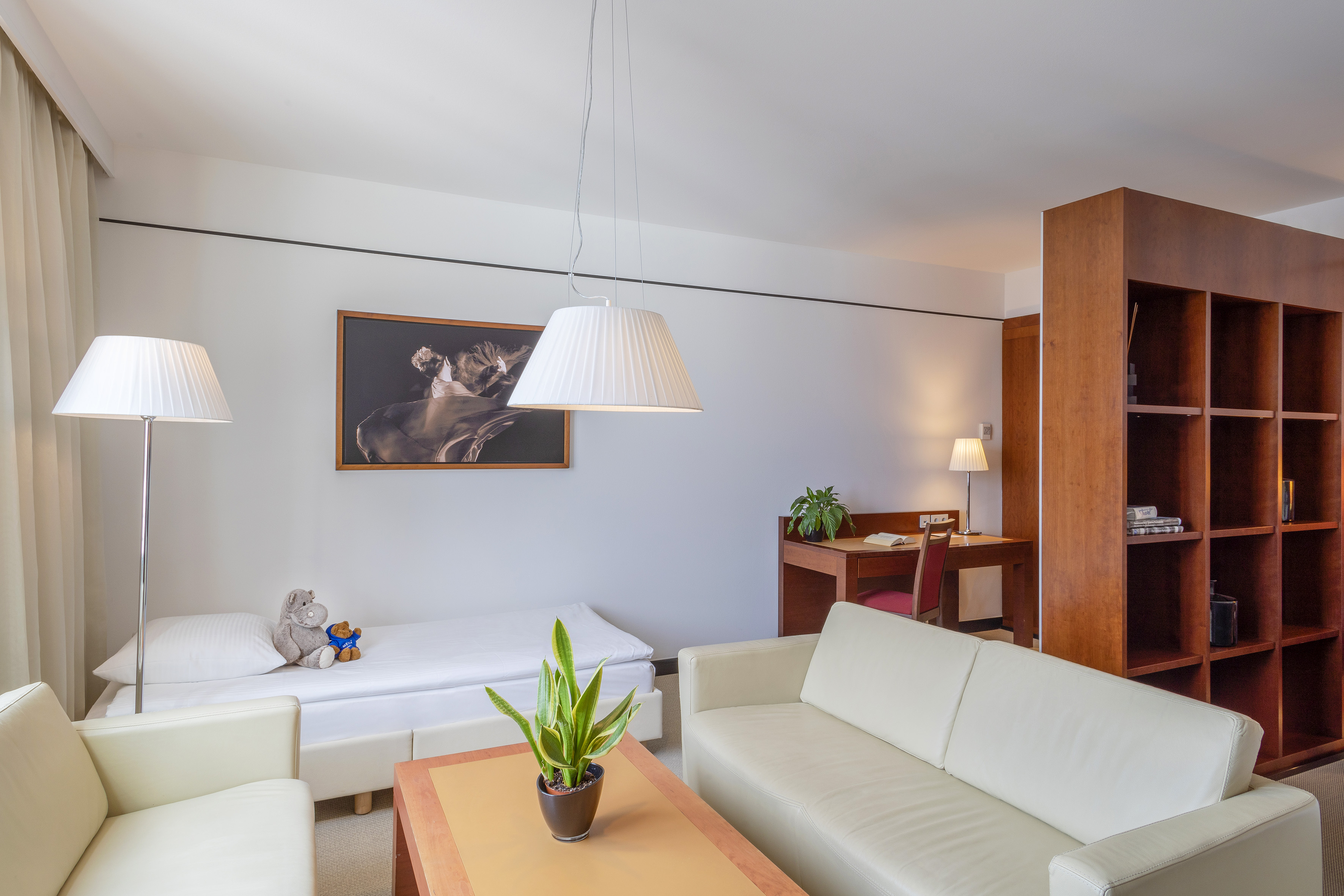 Modern hotel suite with white sofa, single bed, desk, and wooden bookshelf.