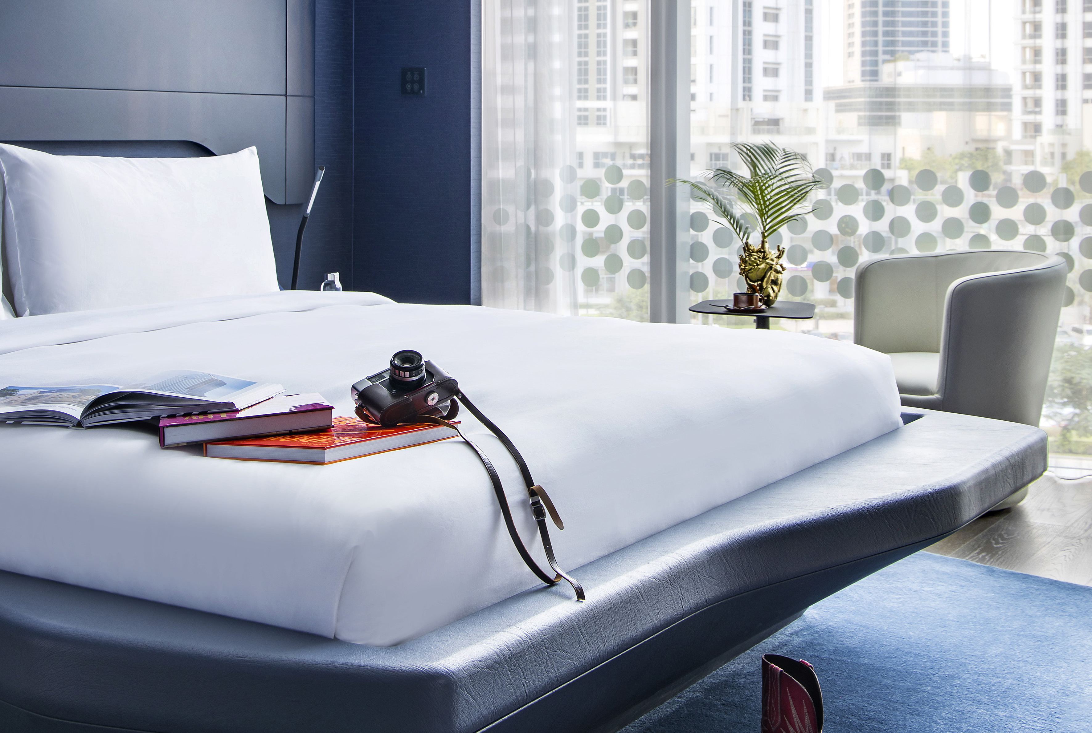 Modern hotel bedroom, white bed, vintage camera, books, city view window.