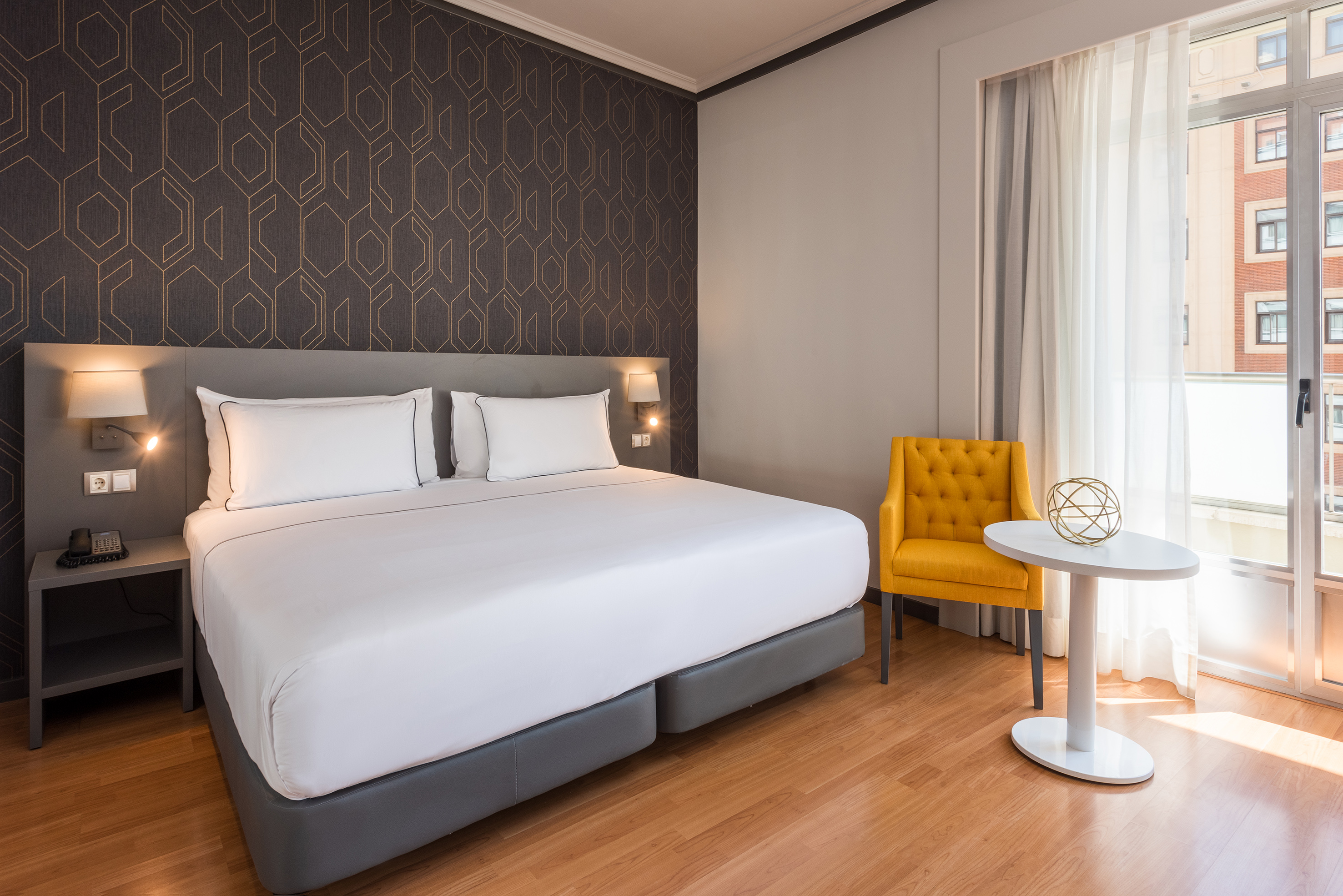 Modern hotel bedroom with king bed, geometric wallpaper, yellow armchair, and window.