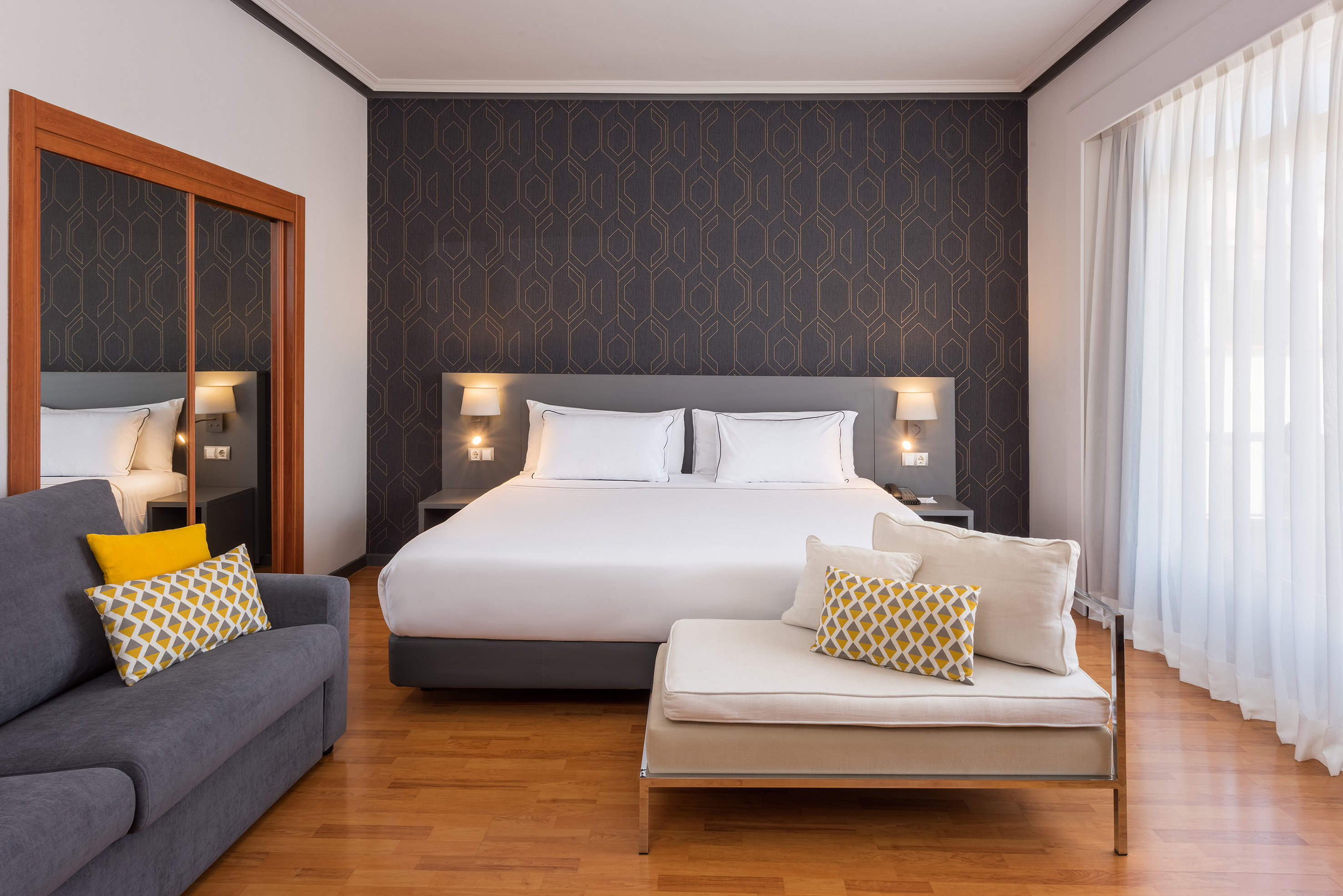 Modern hotel room with king bed, sofa, chaise, and geometric accent wall.