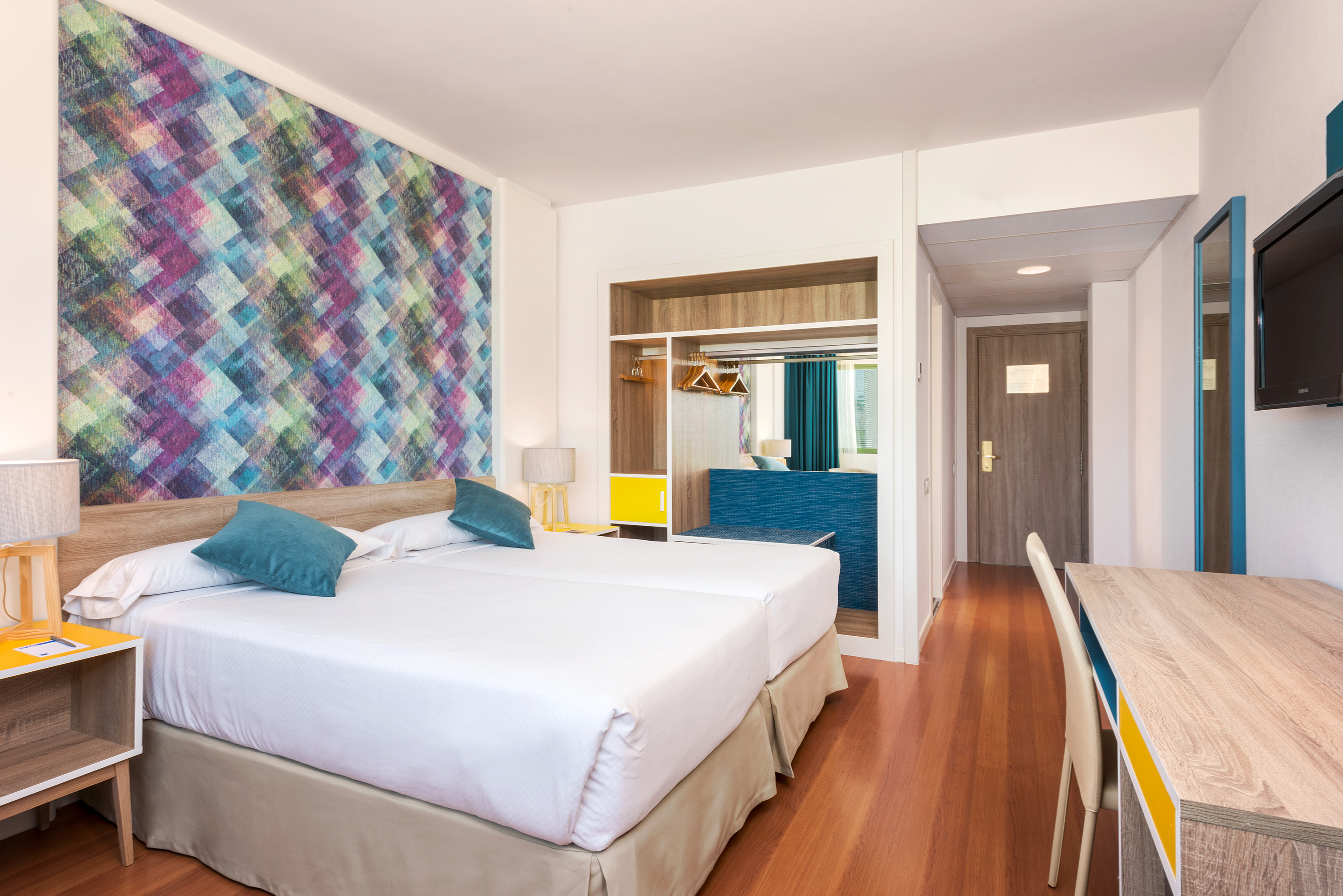 Modern hotel room with vibrant geometric wallpaper, twin beds, and a desk.
