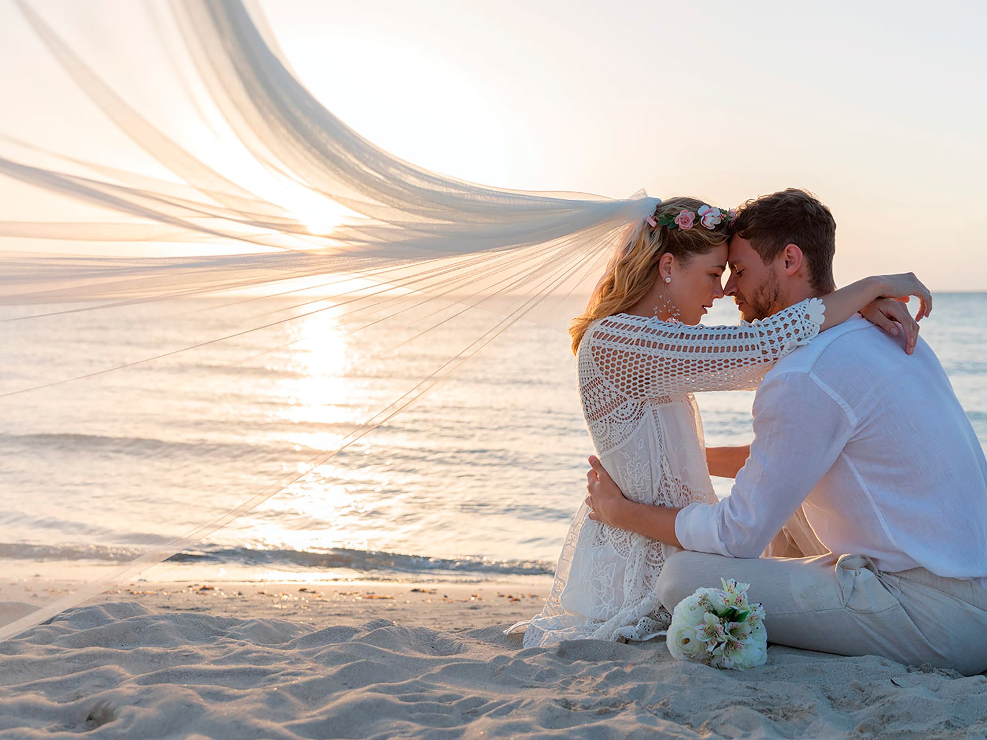 a man and woman sitting on a beach with a veil over their head