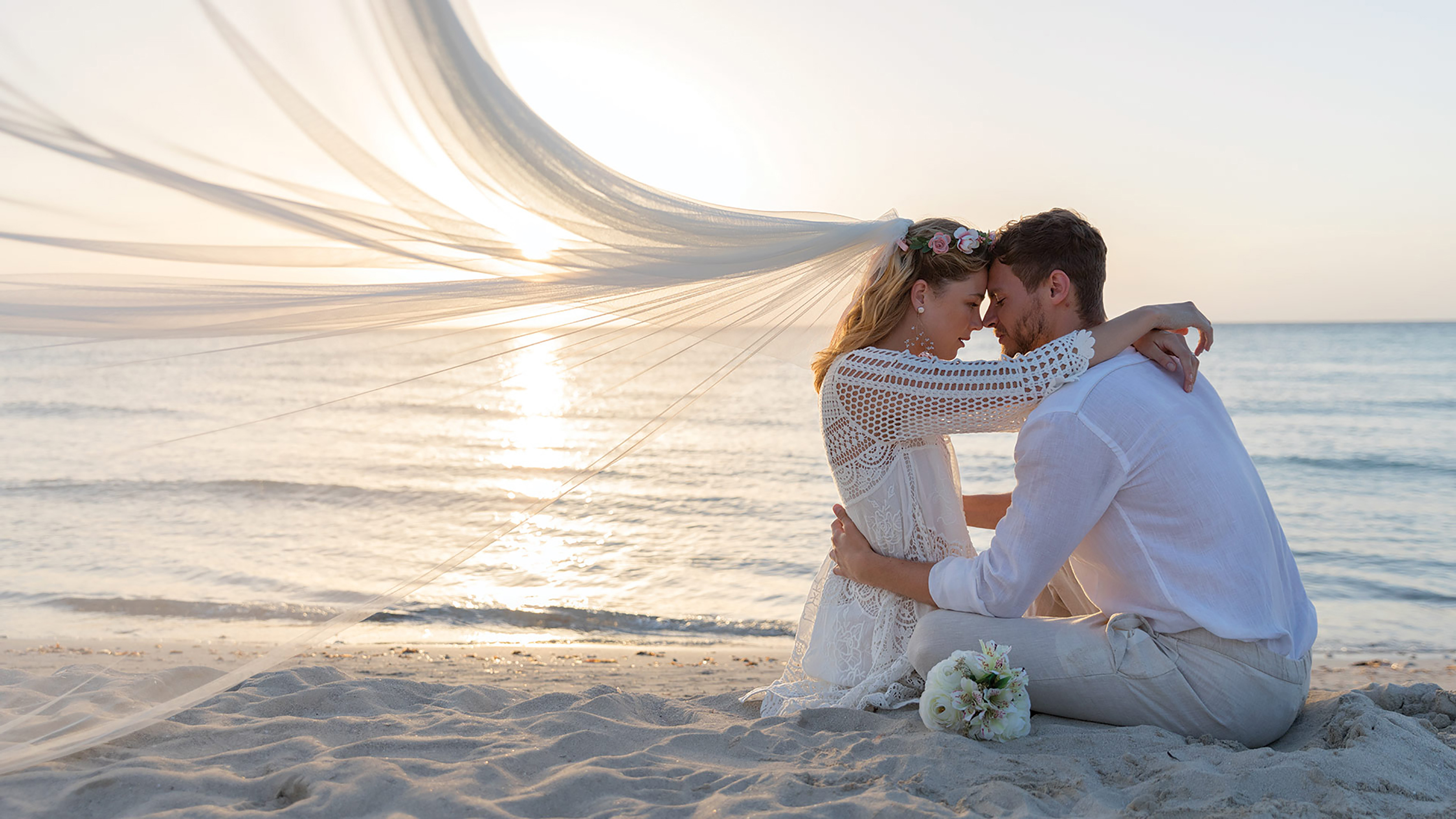 a man and woman sitting on a beach with a veil over their head