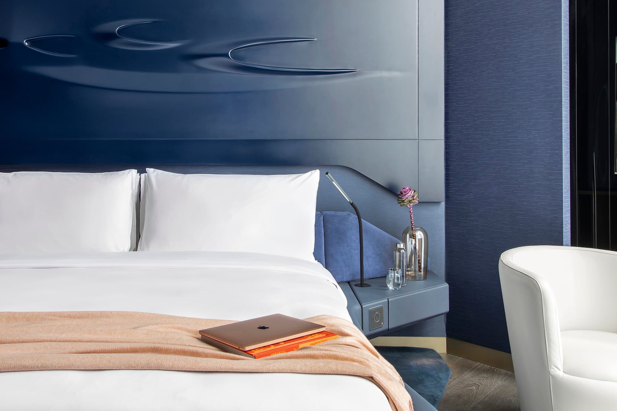 Modern hotel bedroom with navy blue headboard, white bed, laptop, and armchair.