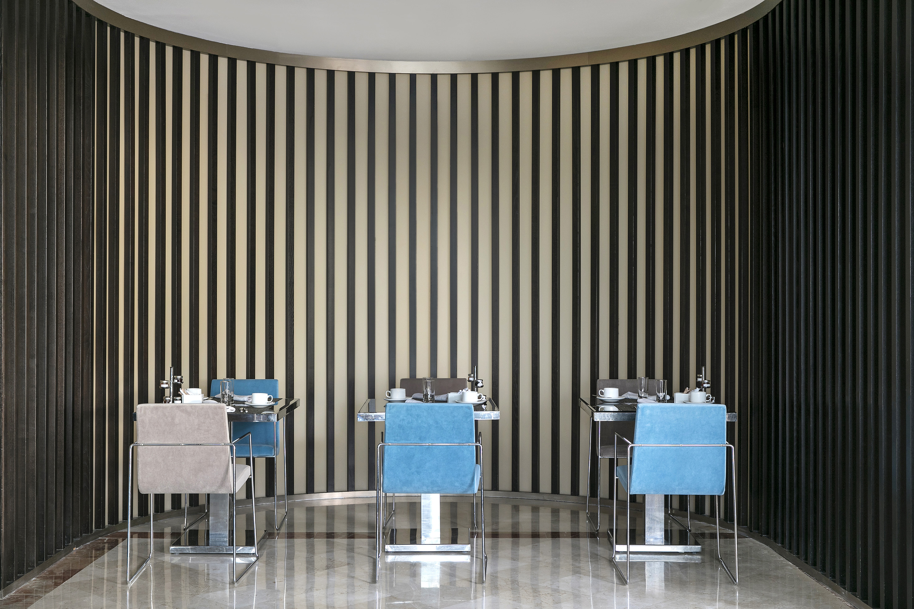 Modern dining area with curved slat wall, blue and beige chairs, set tables.