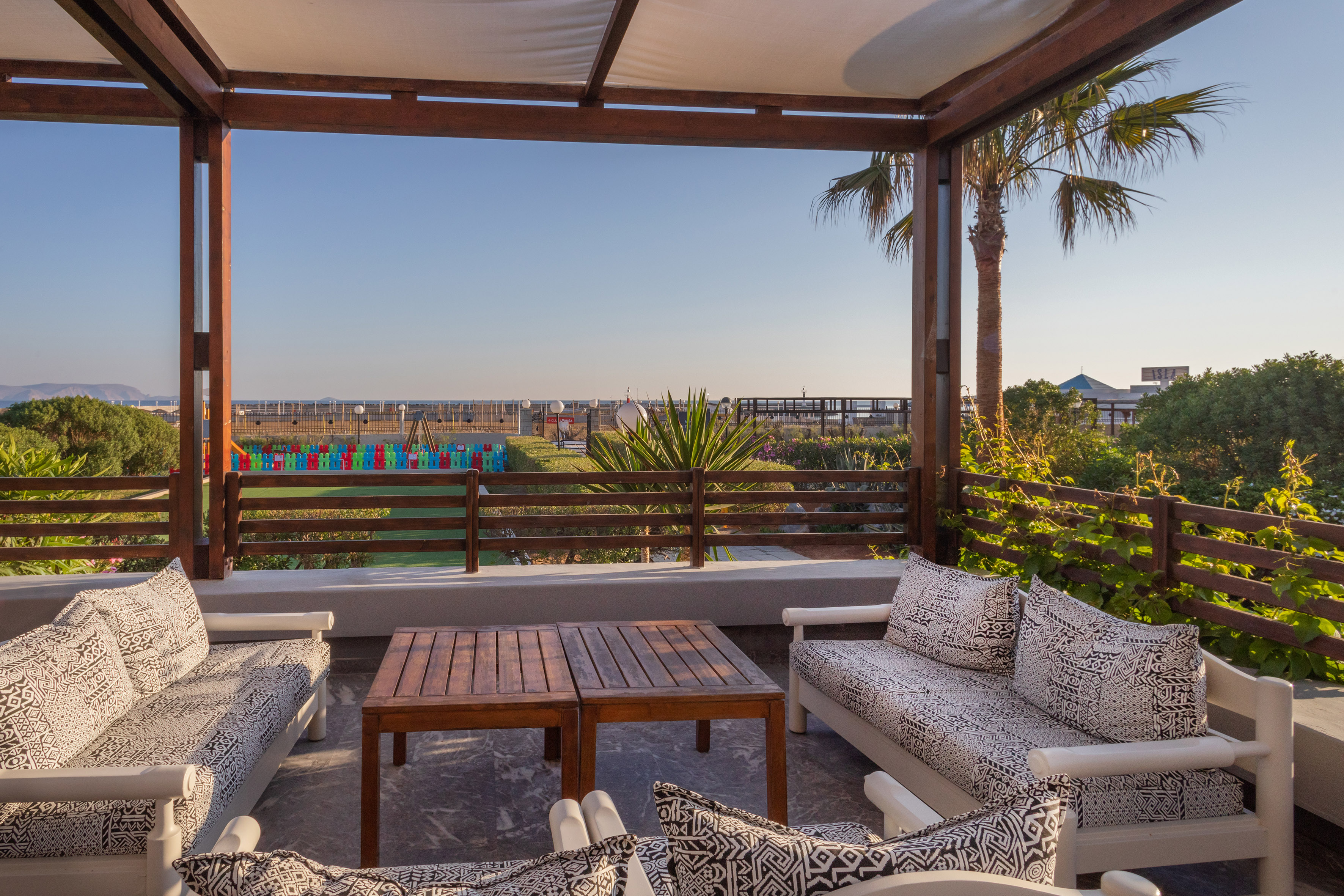Patio with patterned sofas, wooden tables, sea view, and distant play area.