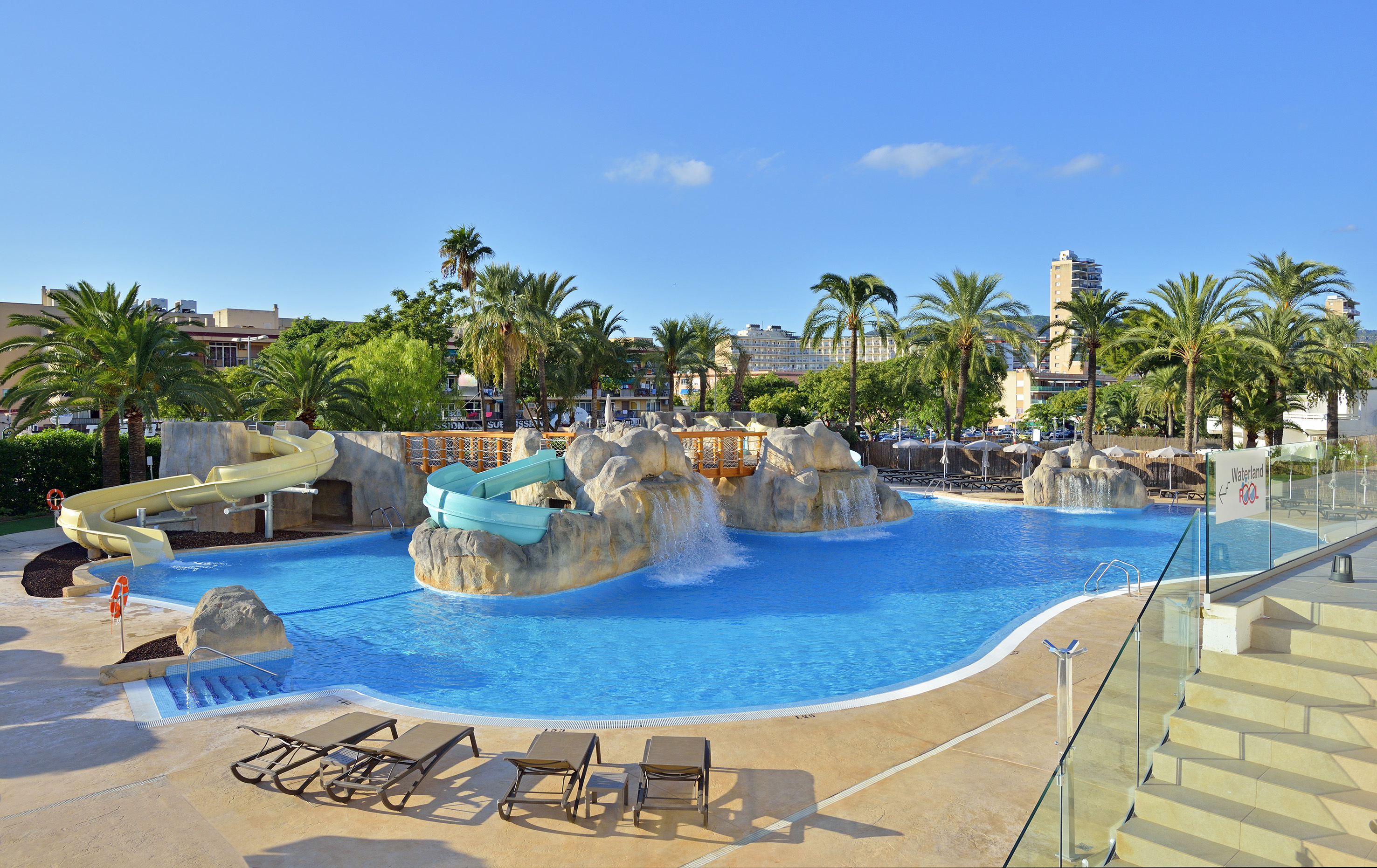 Sunny hotel pool with water slides, waterfalls, and palm trees.