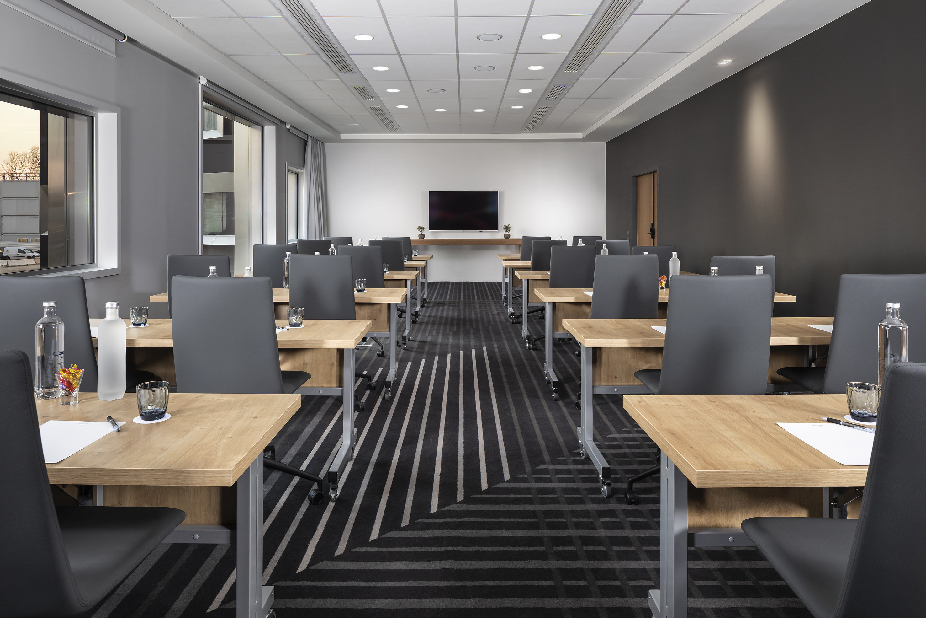 Modern meeting room with individual desks, dark chairs, large screen, and striped carpet.