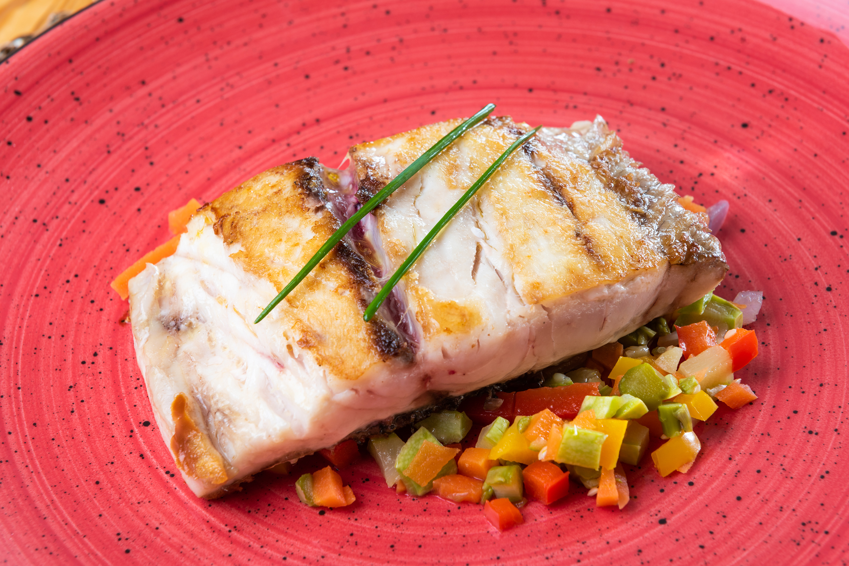 Grilled fish fillet with chives and colorful diced vegetables on a red plate.