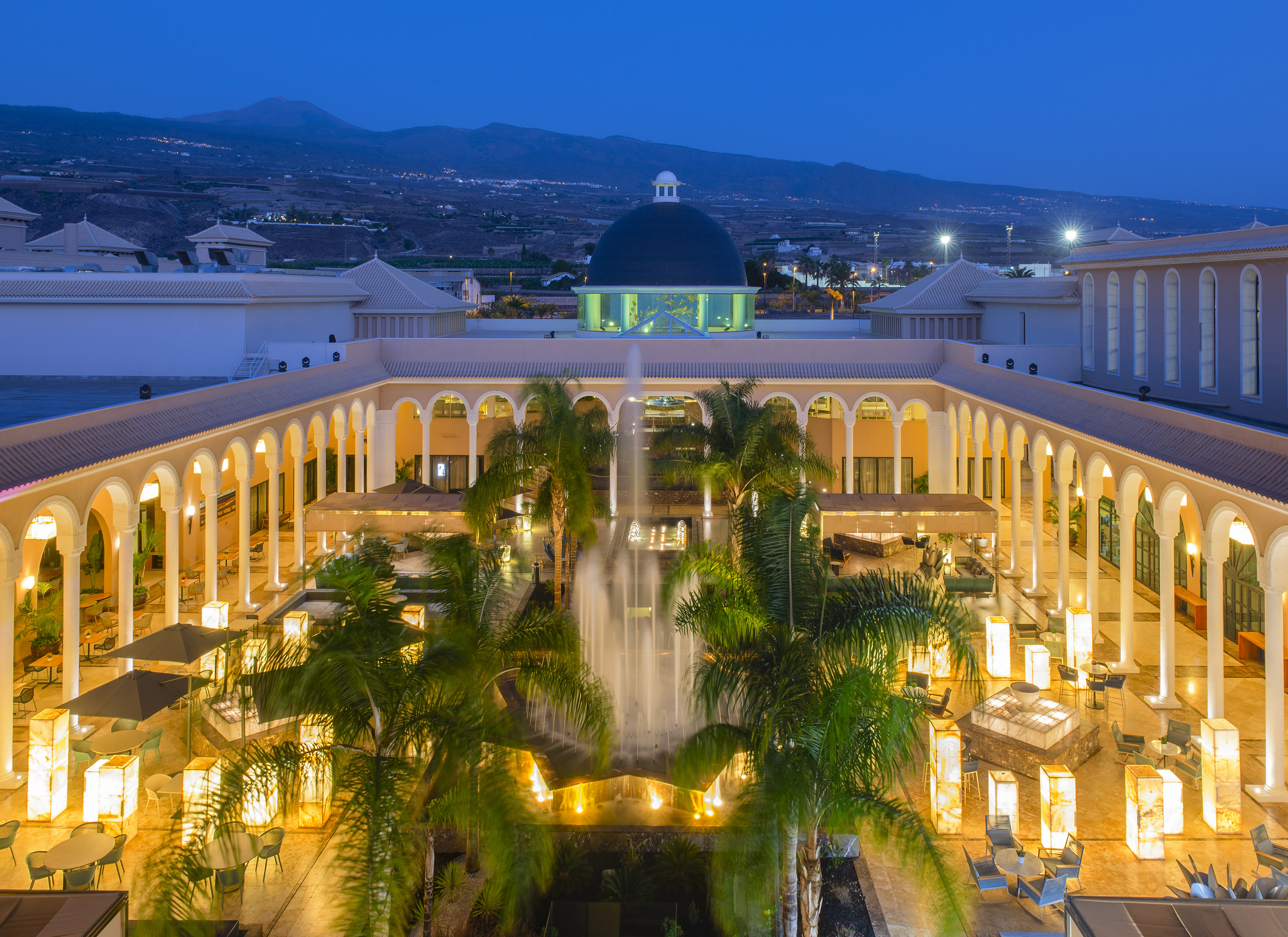 Luxury resort courtyard at night with illuminated fountain, palm trees, and archways.
