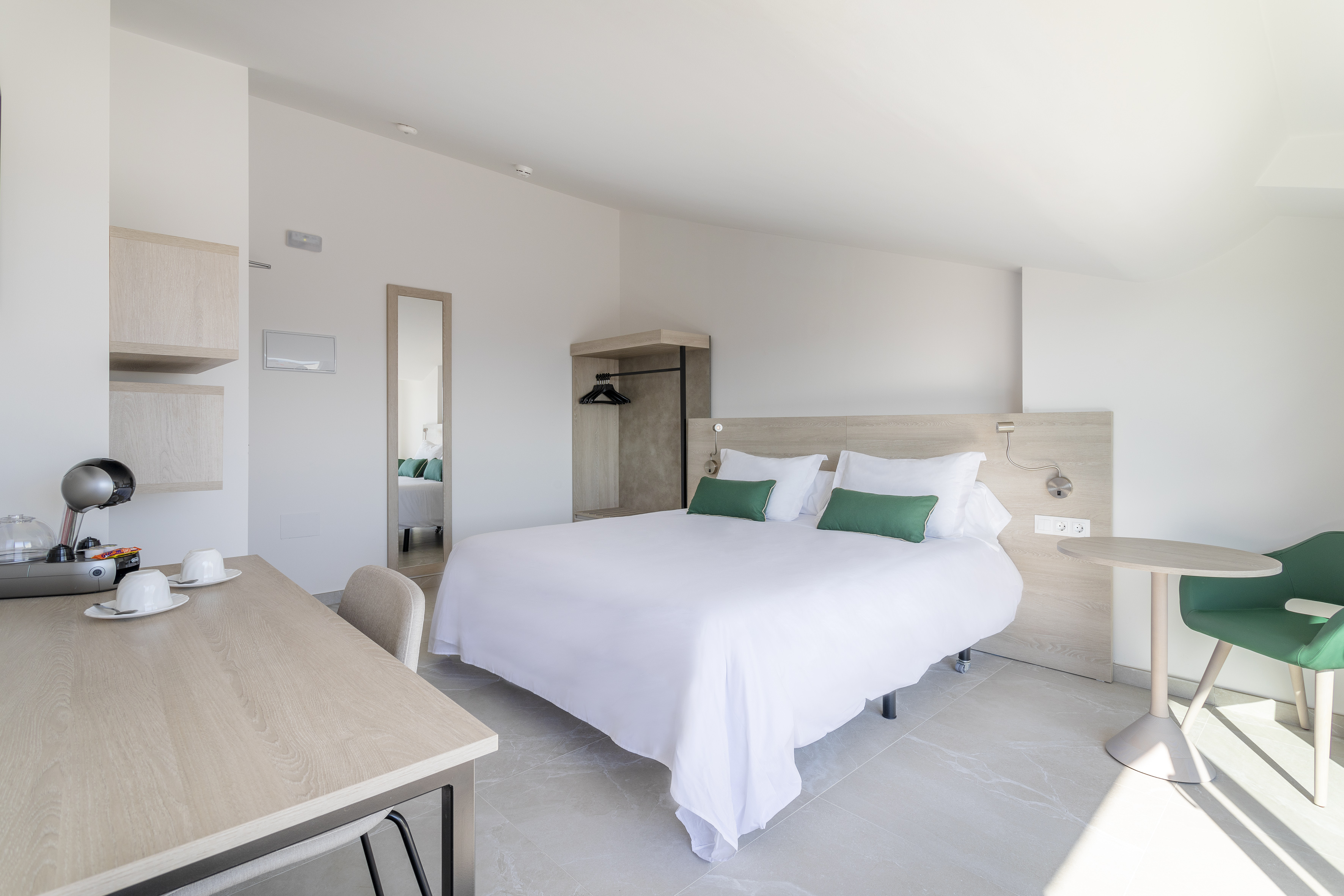 Bright modern hotel room with large bed, green pillows, wooden desk, and coffee maker.
