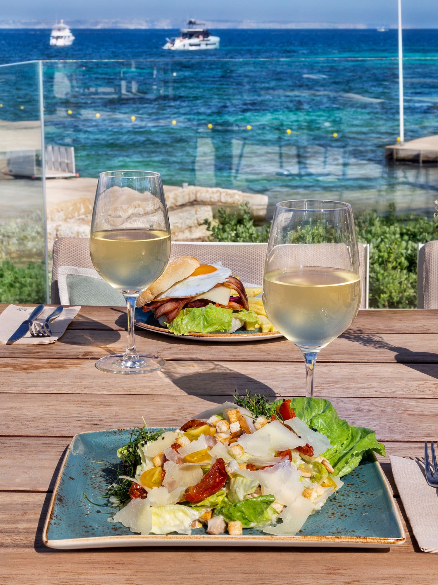 a plate of salad and wine glasses on a table with a body of water in the background
