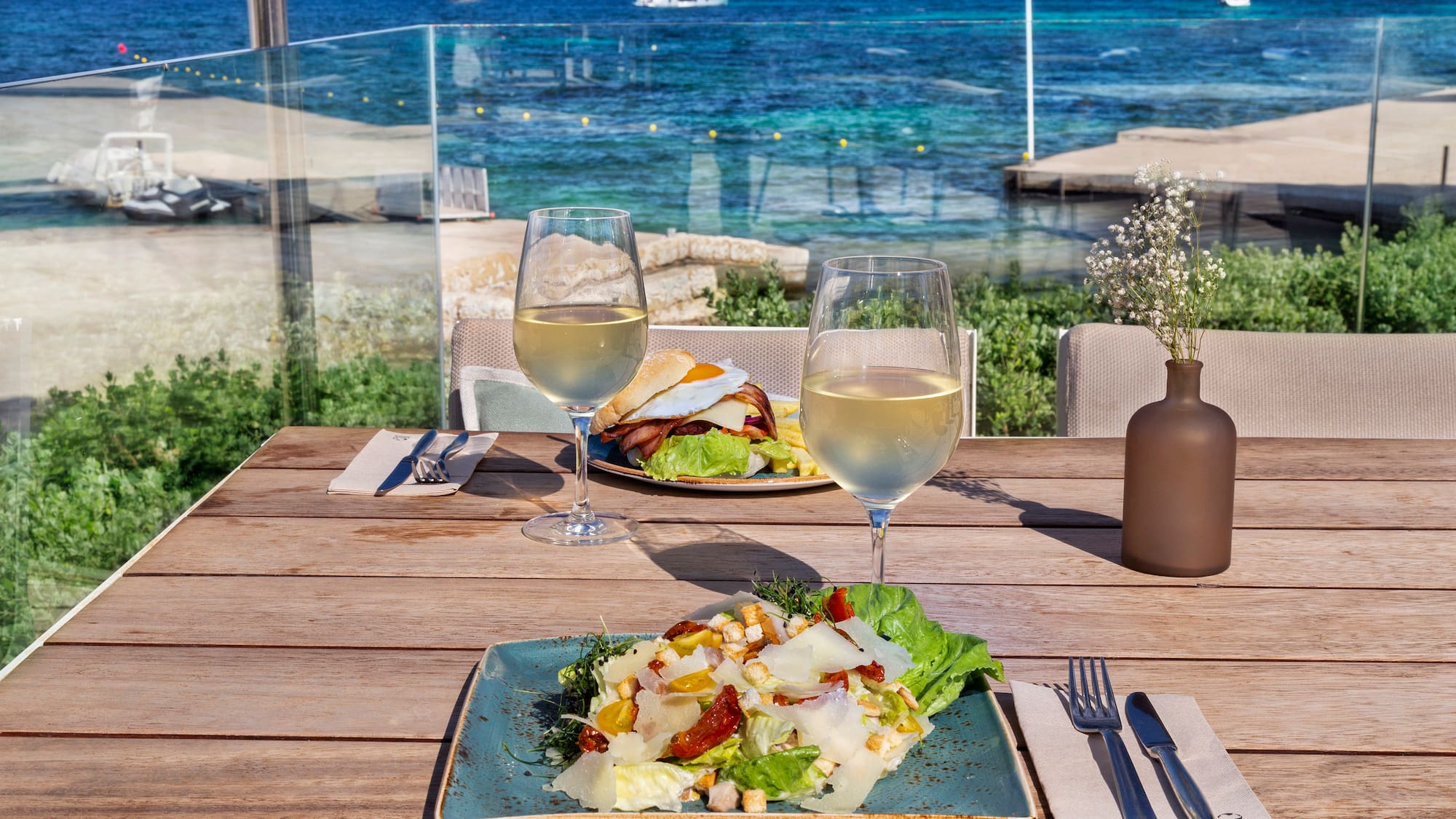 a plate of salad and wine glasses on a table with a body of water in the background