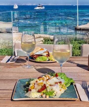 a plate of salad and wine glasses on a table with a body of water in the background