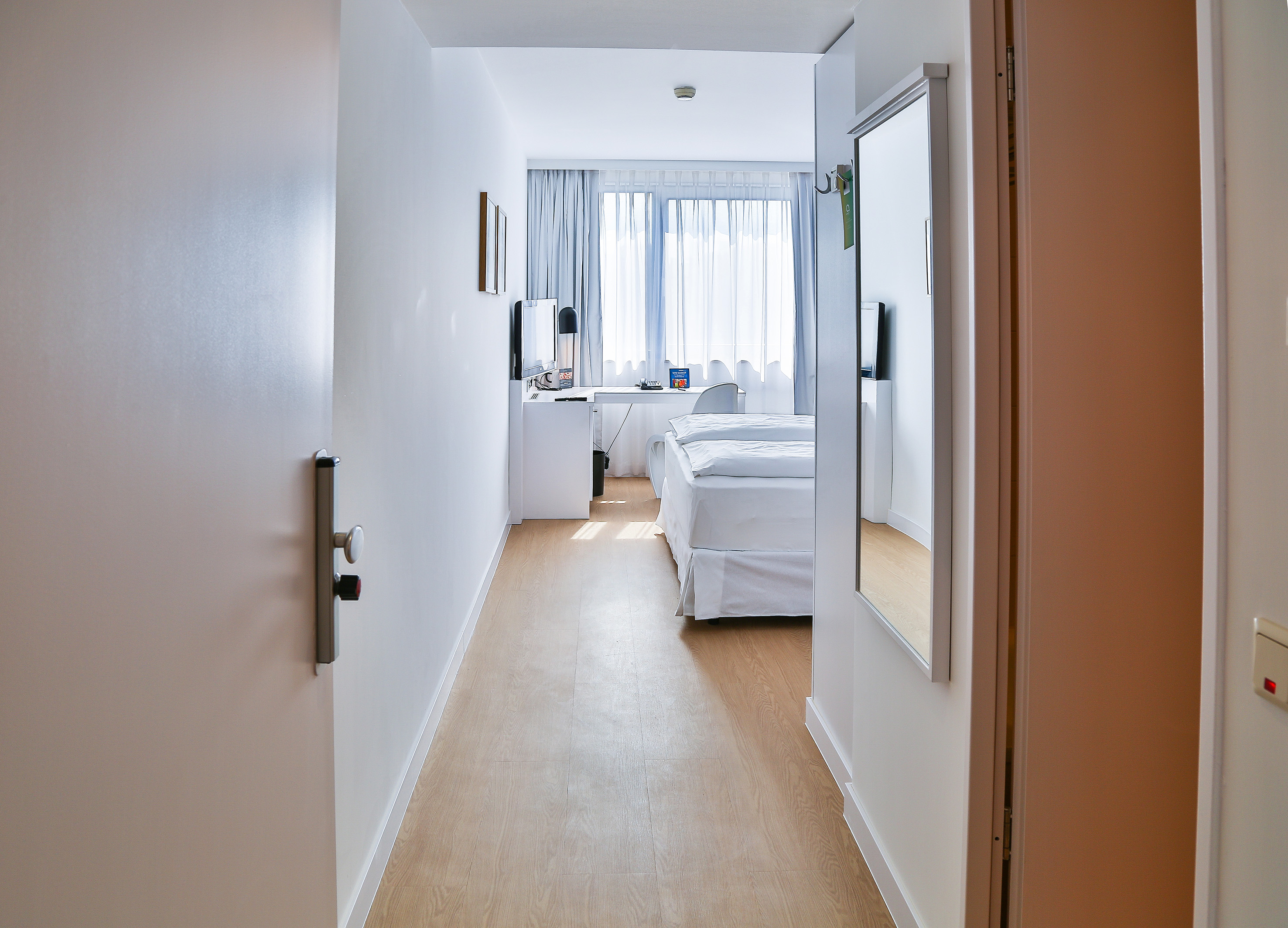 Modern hotel room with twin beds, desk, bright window, and wooden floor.